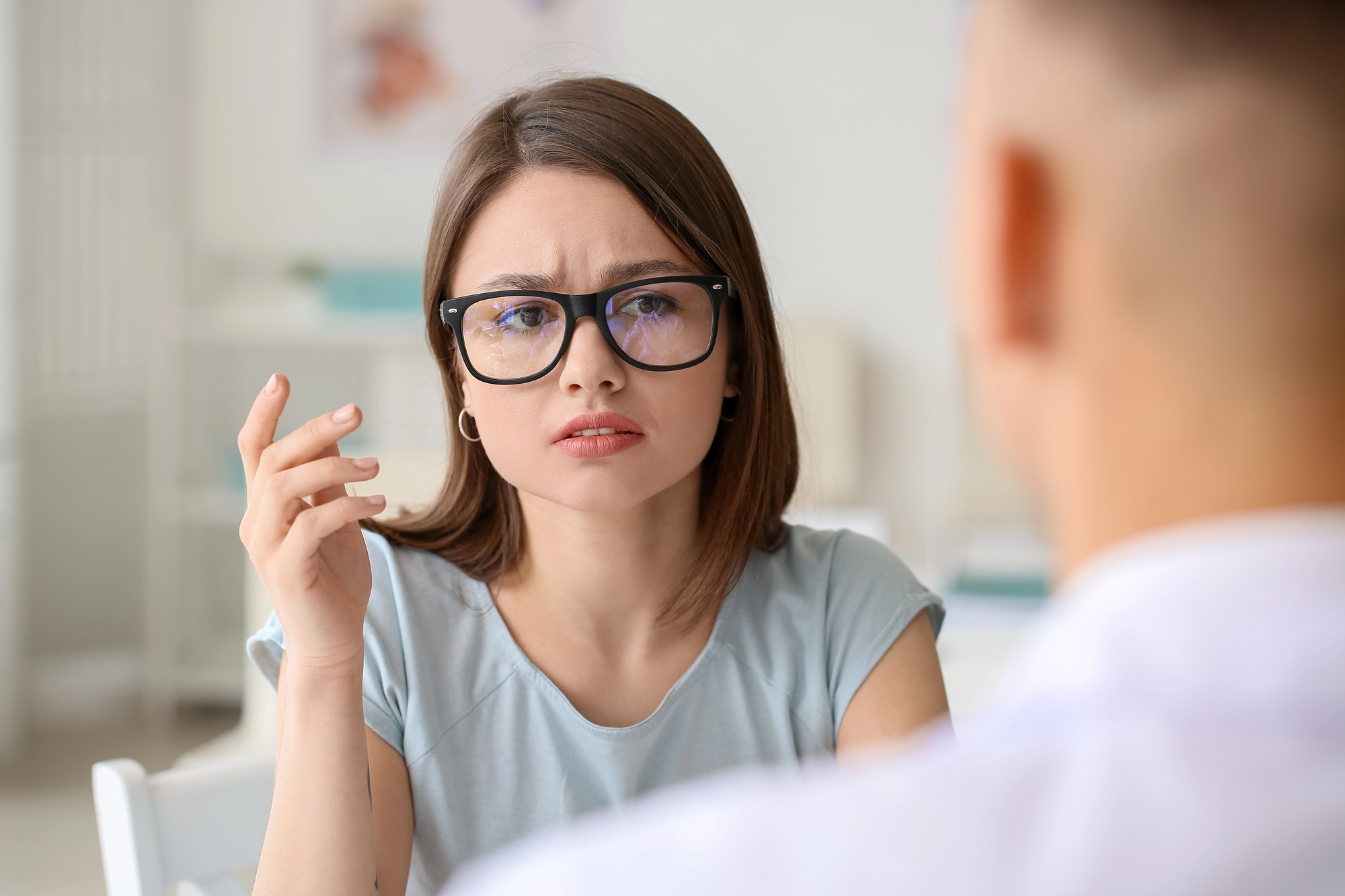 A woman with glasses and a focused expression is sitting and gesturing with her hand, looking at a man whose back is to the camera. The setting appears to be an office or meeting room.