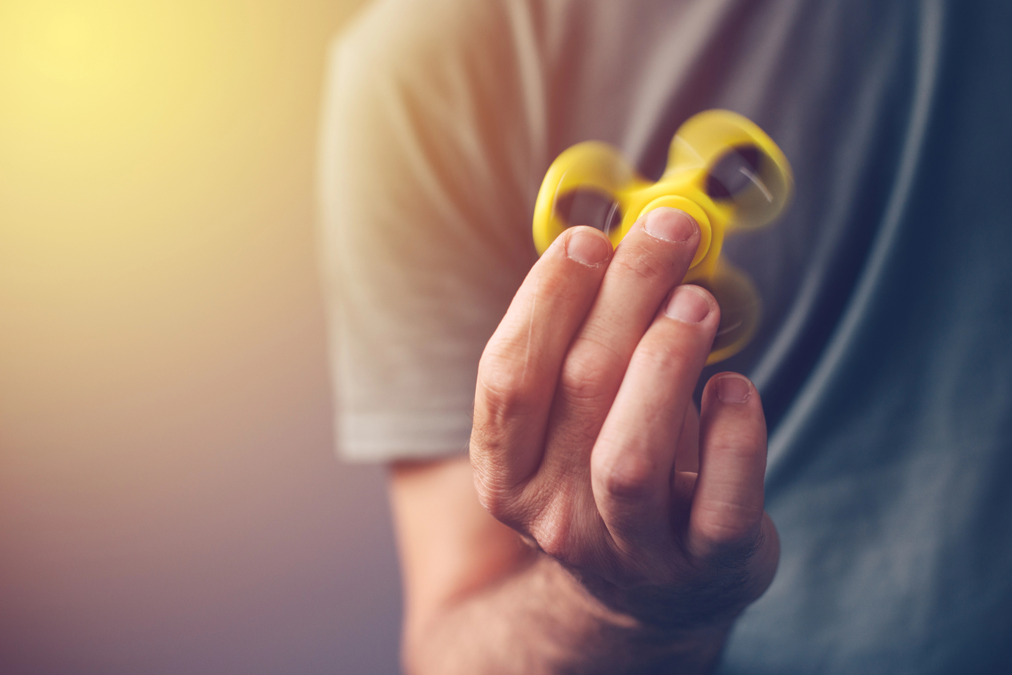 A person in a gray shirt holding a spinning yellow fidget spinner against a blurred background with warm lighting.