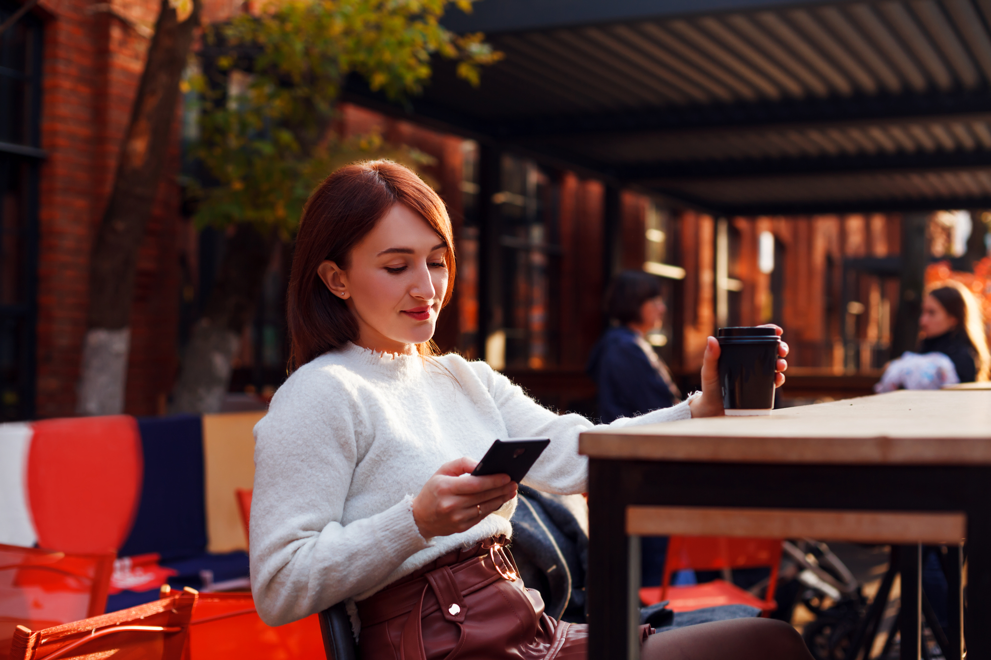 A woman with shoulder-length hair, dressed in a white sweater and sitting at a wooden outdoor table, looks at her phone while holding a black coffee cup. The background features red chairs and a shaded area with trees.