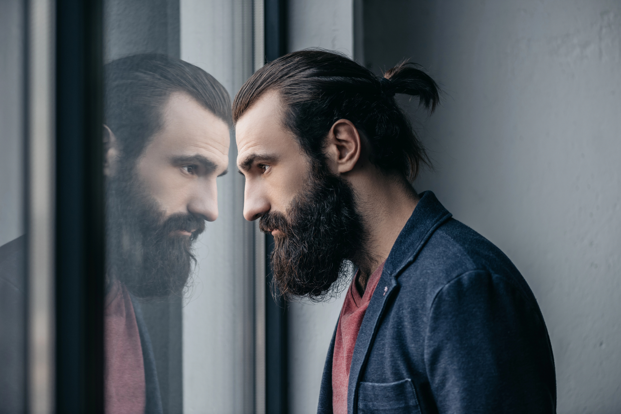 A bearded man with a ponytail is standing indoors, looking thoughtfully out a window. His reflection is visible on the glass. He is wearing a dark blazer over a red shirt. The lighting is soft and neutral.