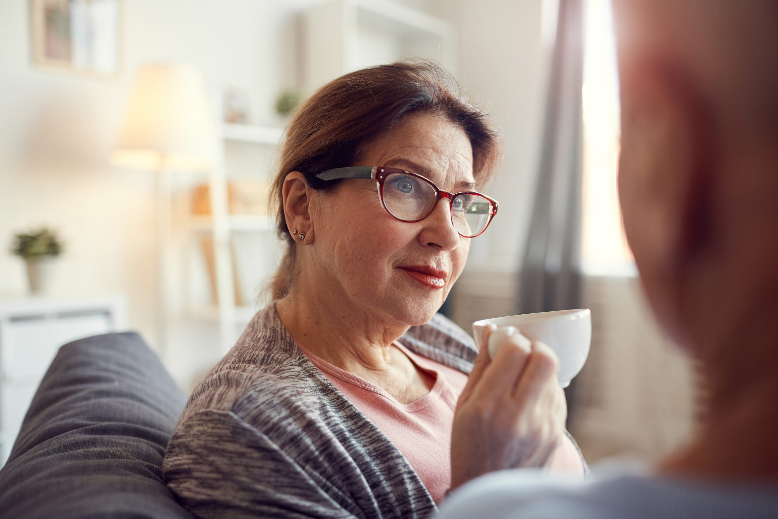 A woman with glasses and a thoughtful expression is sitting on a couch, holding a white teacup. She is indoors in a cozy, well-lit living room with soft furnishings and a lamp in the background.