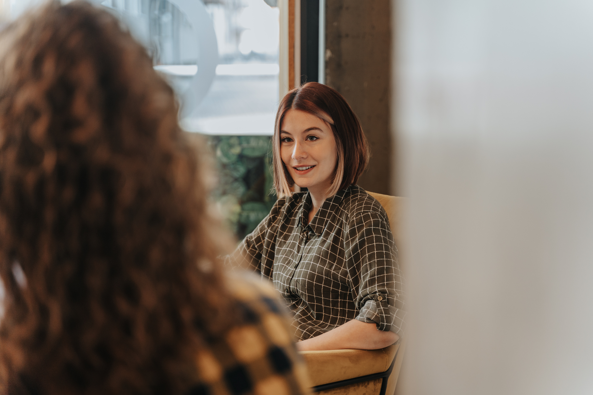 A woman with shoulder-length hair and a checkered shirt is smiling and seated at a table. Her chair is next to a large window. Another person, slightly out of focus, is sitting across from her. The setting appears to be casual or work-related.