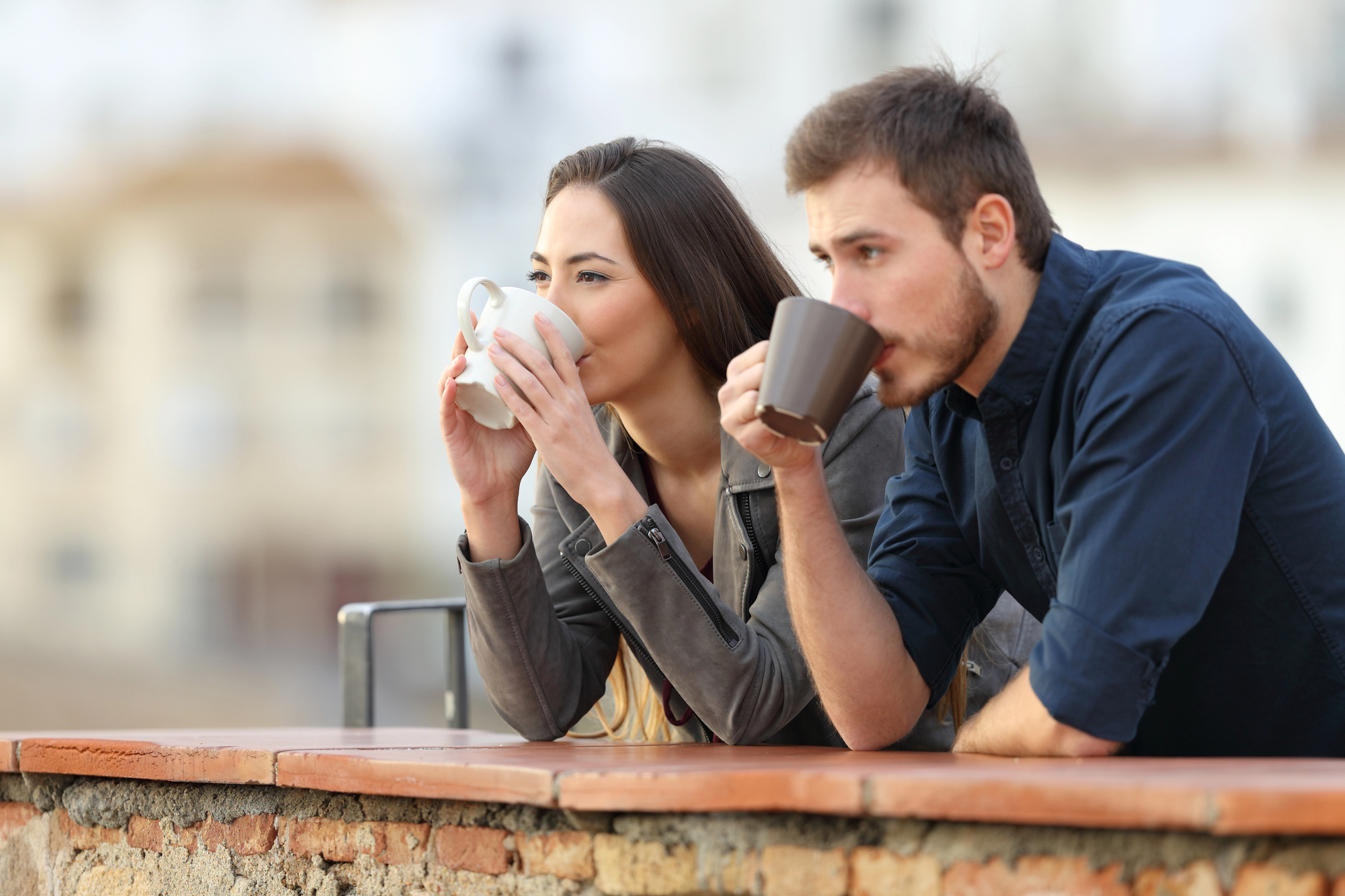 A man and woman lean on a balcony railing, sipping from mugs. They are dressed casually and appear relaxed, enjoying their drinks while gazing into the distance. The background is blurred, suggesting an outdoor urban setting.