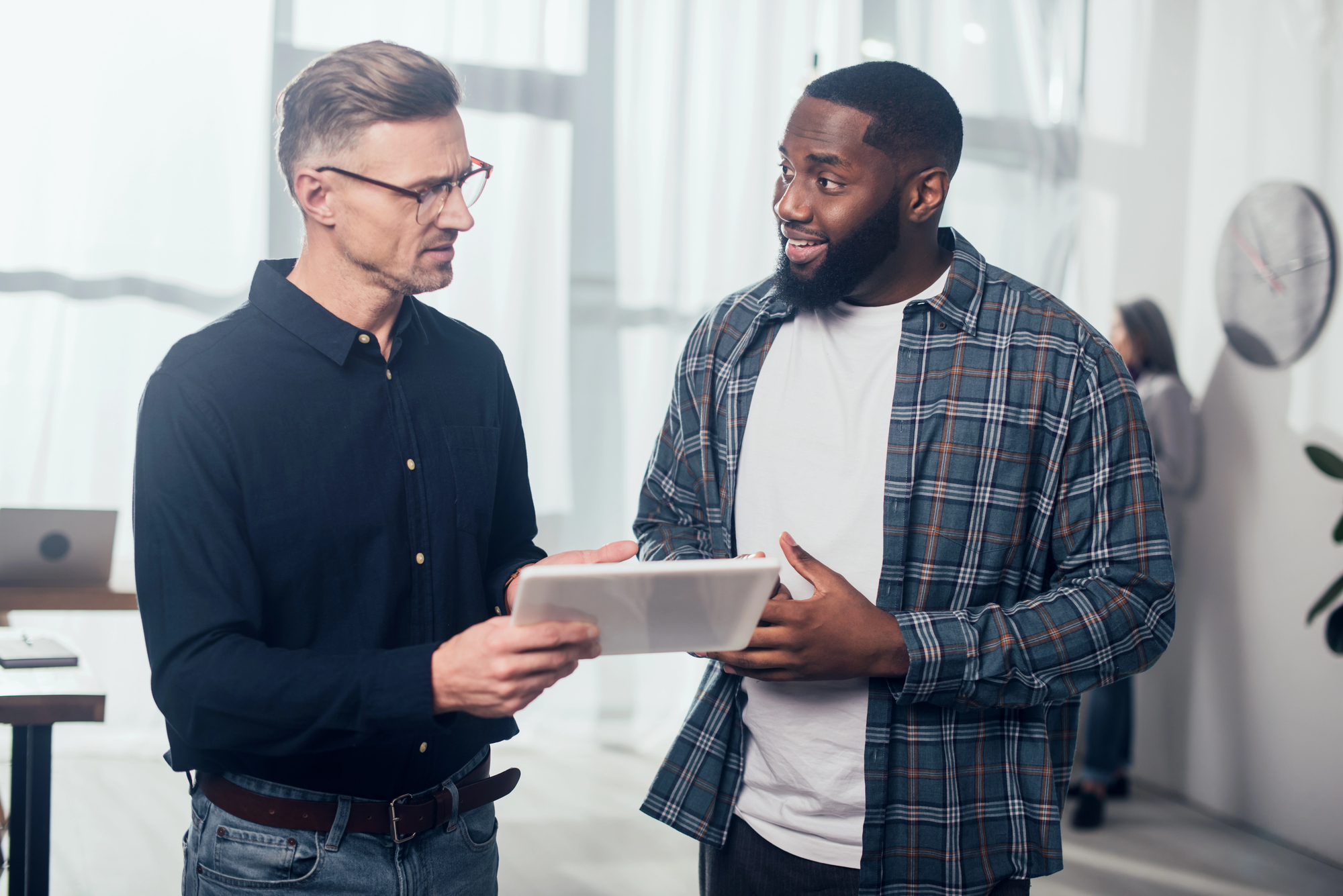 Two men in a bright office are having a discussion. One is holding a tablet, and they both look engaged in conversation. A clock and office furniture are visible in the background.