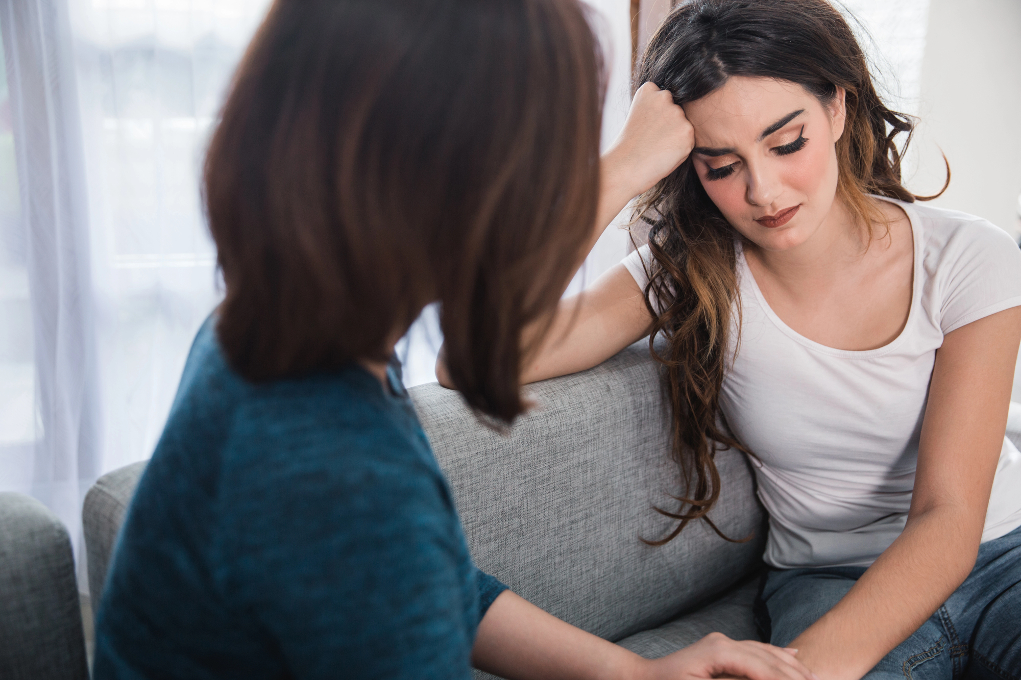 Two women sit on a couch, engaged in a serious conversation. The woman on the right looks visibly upset, resting her head on her hand. The woman on the left seems to be comforting her, with a supportive gesture. They are in a bright room.