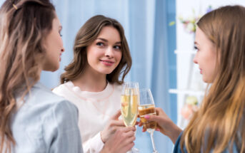 Three women smiling and clinking champagne glasses in a celebratory manner, standing indoors with a soft blue background.