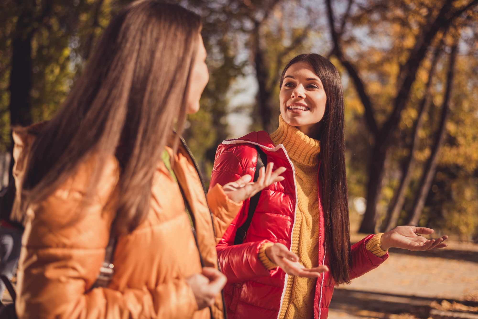 Two women in colorful jackets are walking and conversing in a park surrounded by autumn trees. The woman on the right is gesturing with her hands, and both appear to be enjoying their conversation.