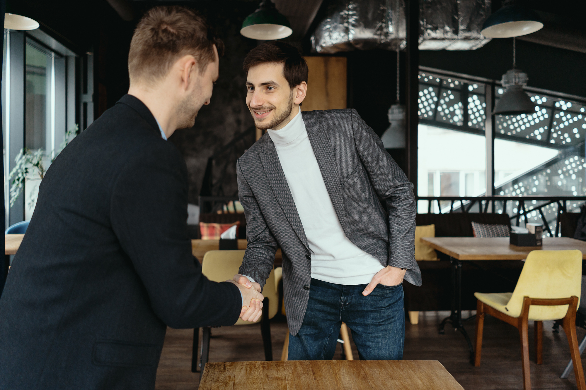 Two men in a modern office setting are shaking hands across a wooden table. One is wearing a gray blazer with a white turtleneck, and the other is in a dark suit. They appear to be having a friendly meeting or greeting.