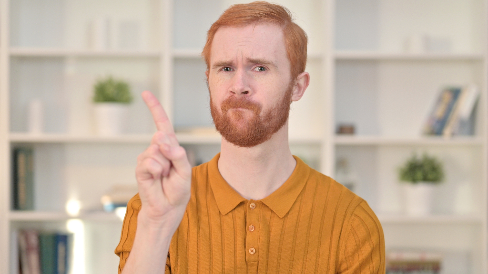 A red-haired man with a beard wearing an orange shirt makes a serious face while holding up his index finger. He stands in front of a white bookshelf with plants and books.