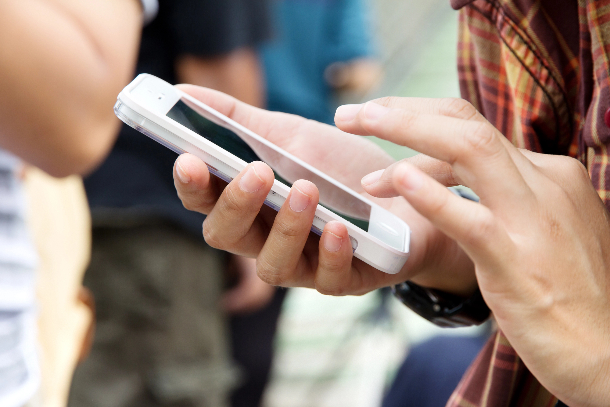 Close-up of a person using a smartphone. The individual is holding the phone in one hand and tapping the screen with the other. They are wearing a patterned shirt. The background is blurred, showing indistinct figures and colors.