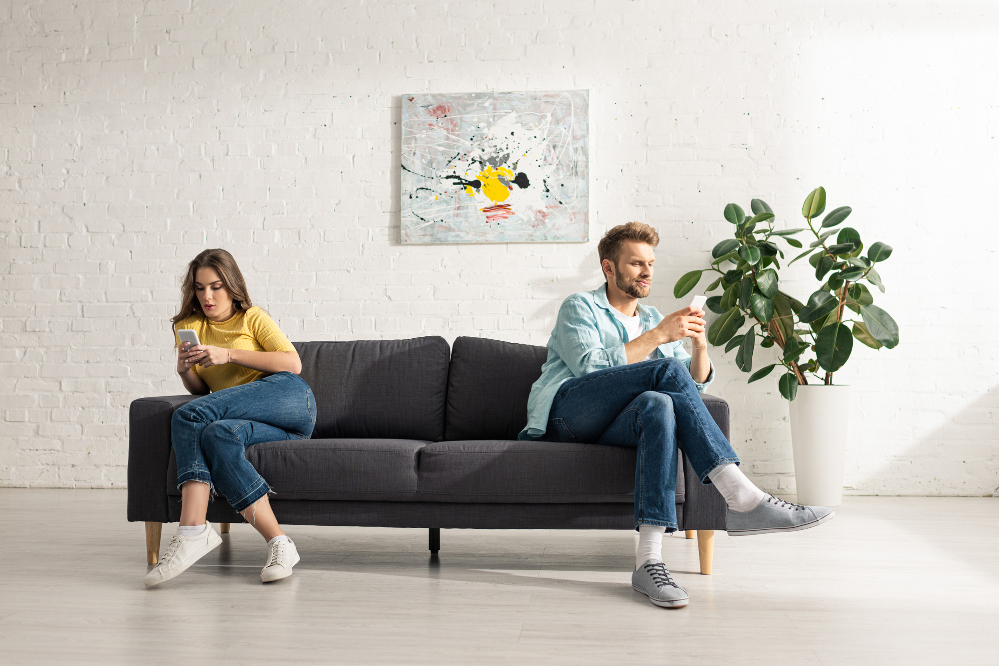 A man and woman sit on opposite ends of a dark gray sofa, both looking at their phones. The woman wears a yellow top and jeans, and the man wears a light blue shirt and jeans. A potted plant and abstract painting decorate the white brick wall behind them.