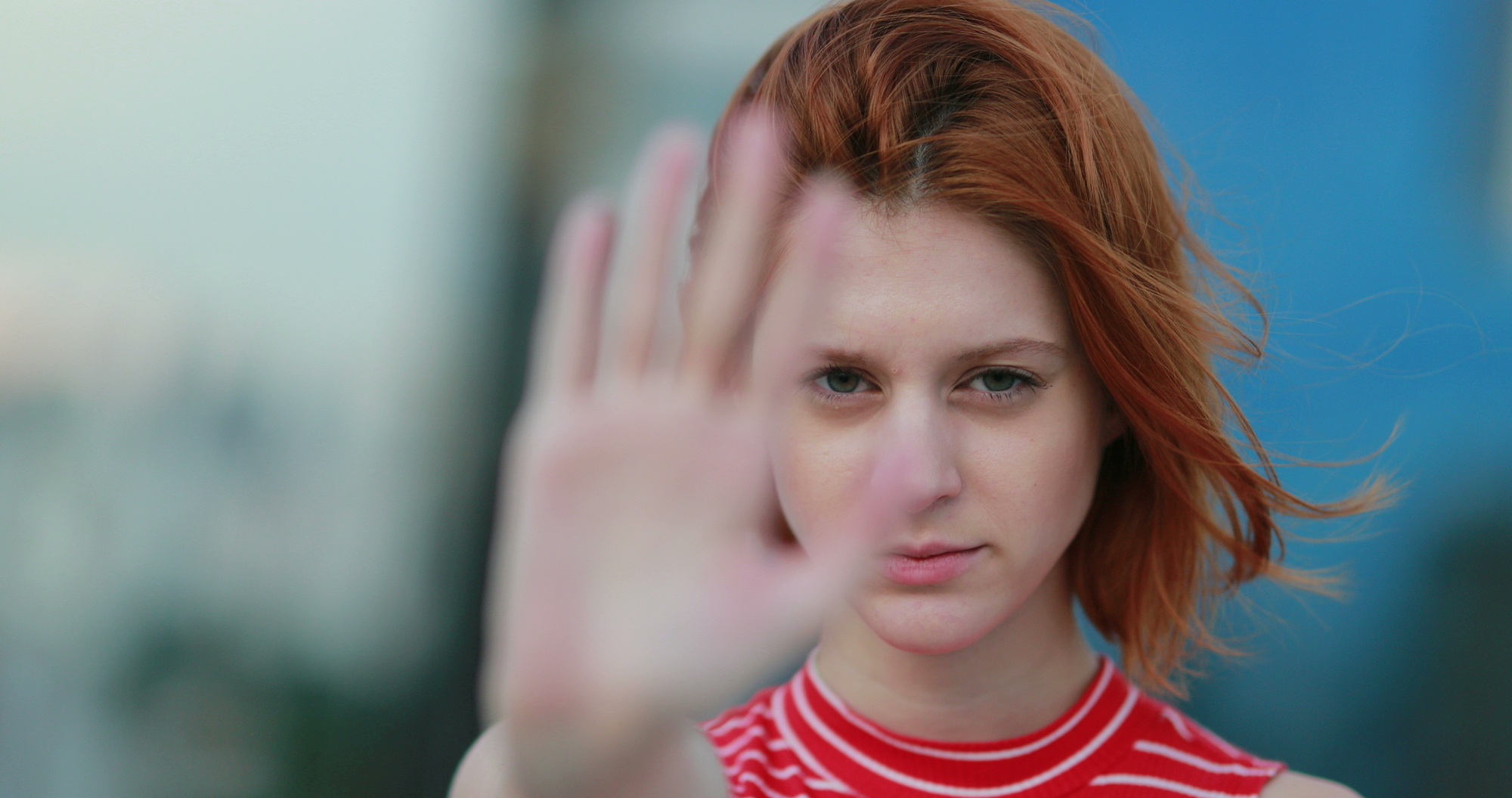 A person with red hair is standing outdoors, wearing a red and white striped top. They are holding their hand up in front of their face, partially blocking it, with a blurry background.