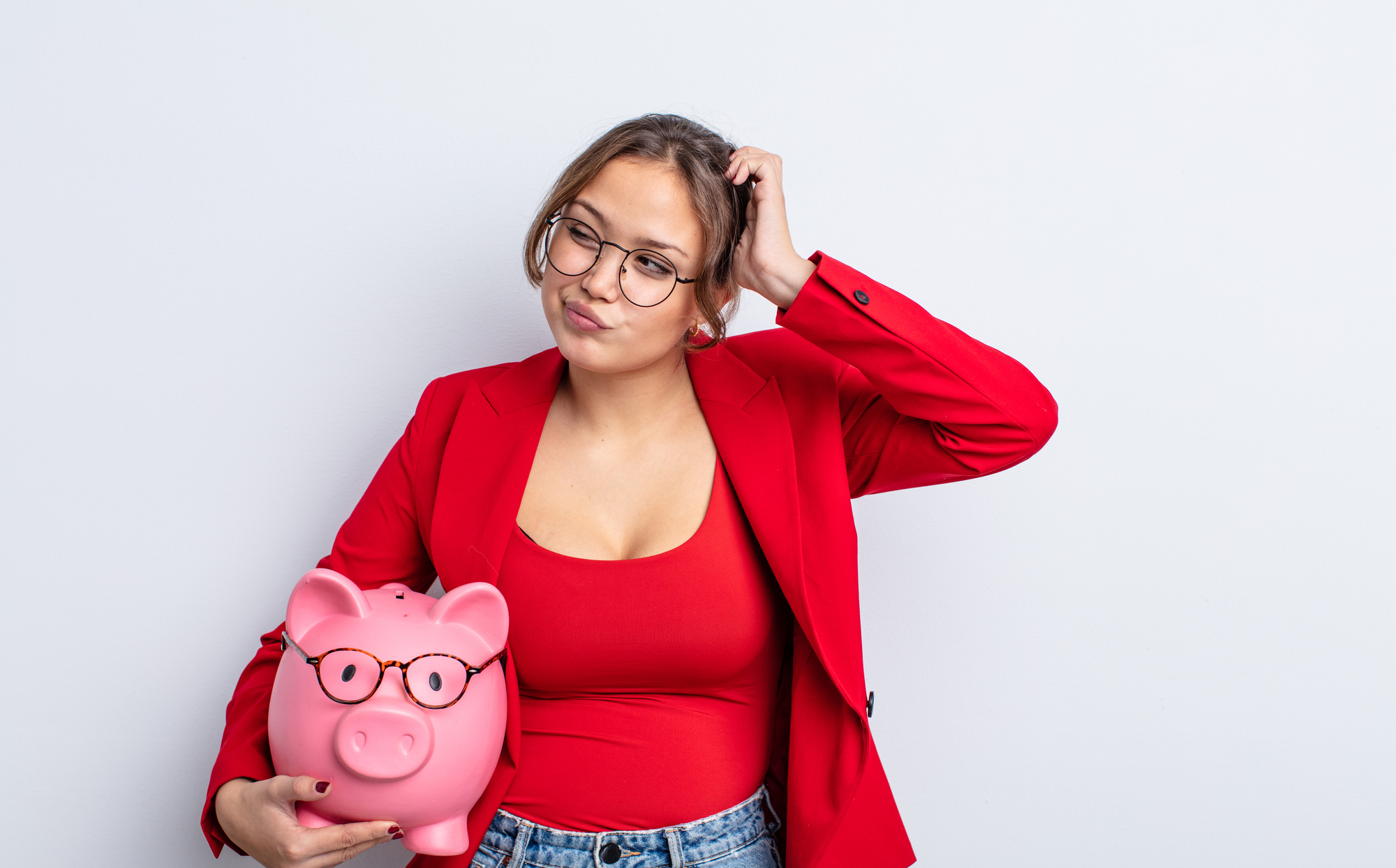 A woman in a red blazer and top, wearing glasses, scratches her head while holding a pink piggy bank also wearing glasses. She stands against a plain white background.