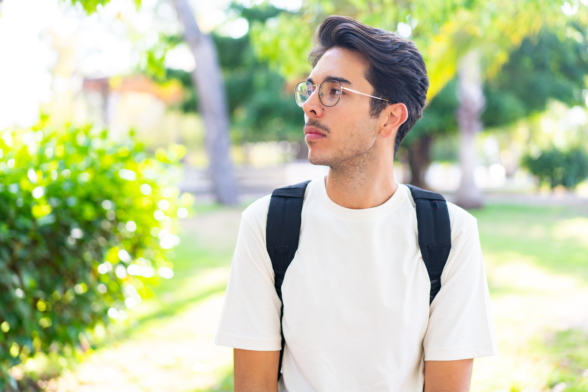 A man wearing glasses and a white shirt stands with a backpack in a park. He looks to his right, surrounded by lush green trees and sunlight filtering through the leaves.