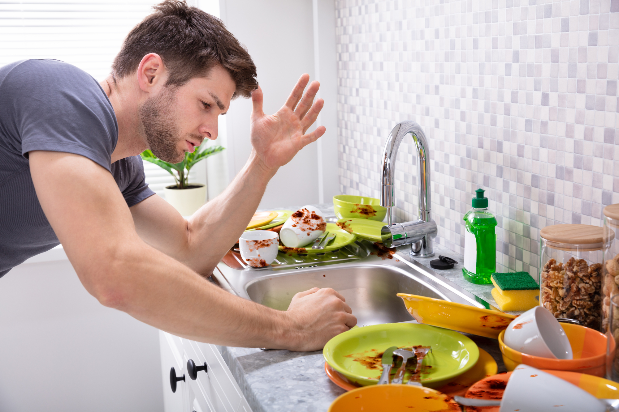 A man in a kitchen is gesturing in frustration at a pile of dirty dishes in the sink. Plates and utensils with food residue are scattered on the counter. A small plant and cleaning supplies are visible in the background.