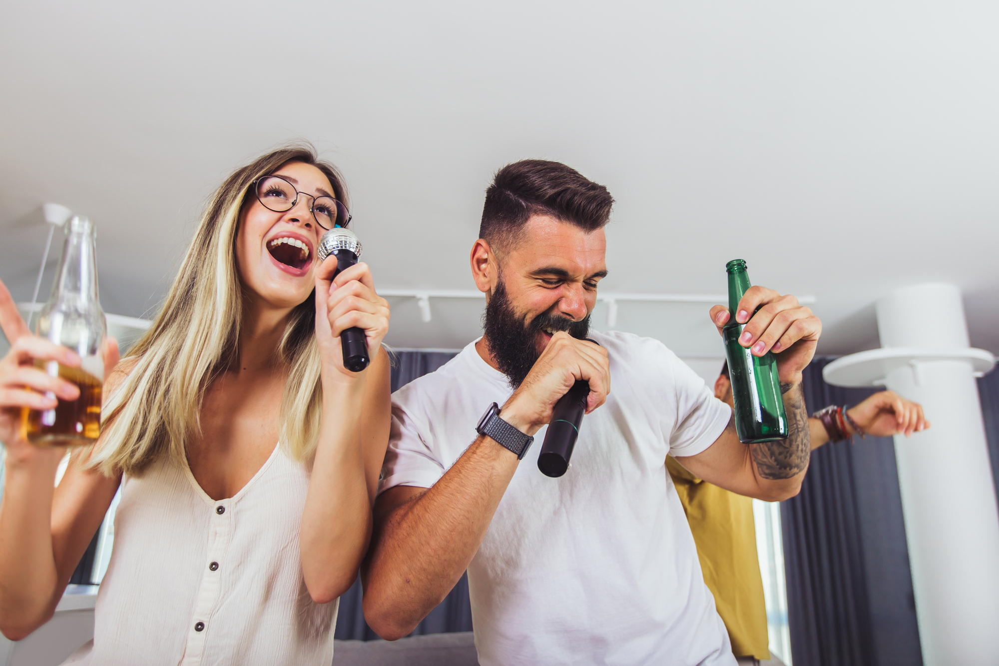 A woman and a man are having fun singing karaoke at home. They are both holding microphones, and each has a drink in hand. The woman is wearing glasses and has long hair, while the man has a beard. Another person is partially visible in the background.