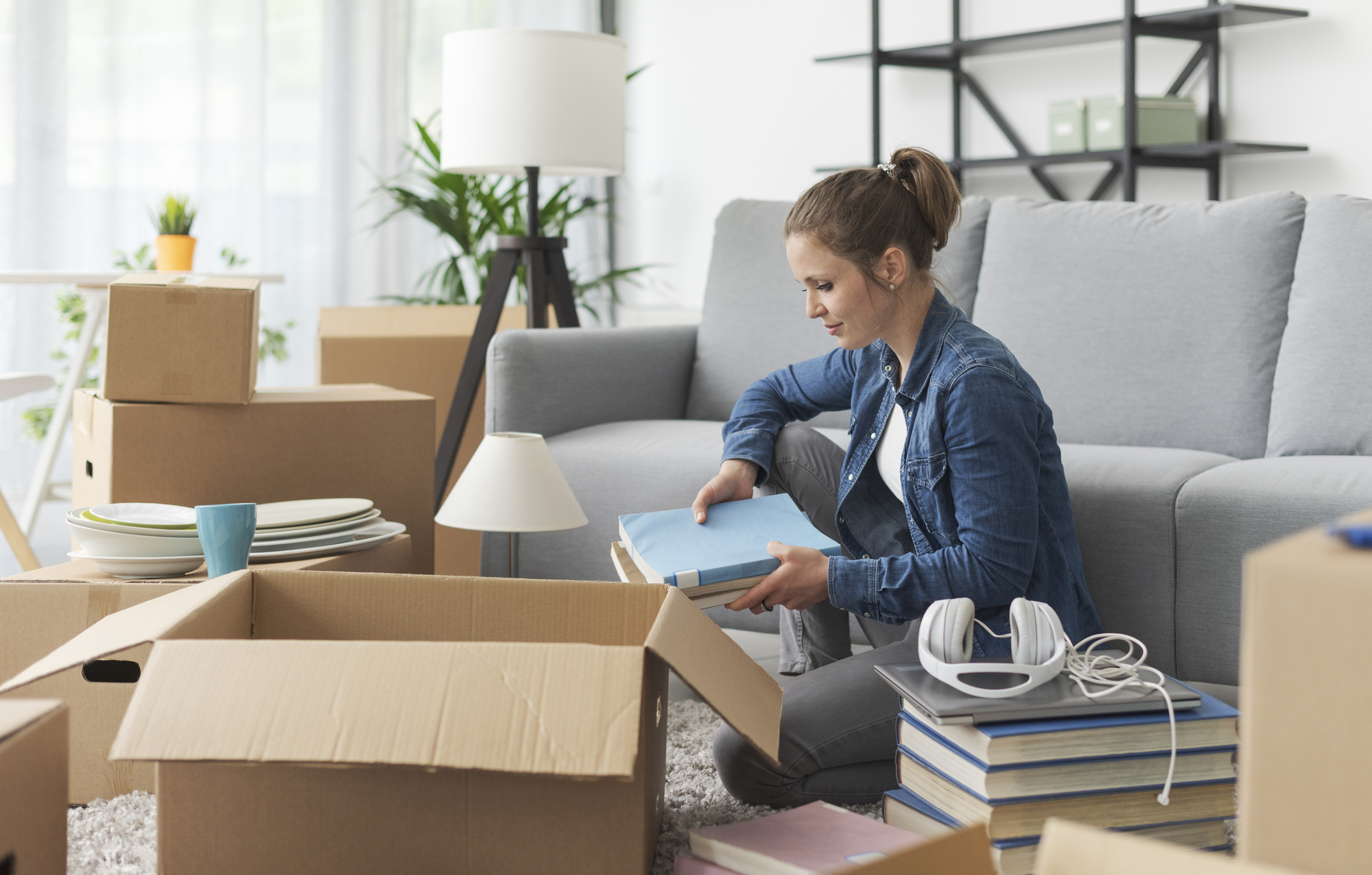 A woman is sitting on the floor in a living room, unpacking boxes. She is placing a book on a stack of books. Surrounding her are cardboard boxes, a lamp, headphones, and a stack of plates. A gray sofa and shelves with books are in the background.