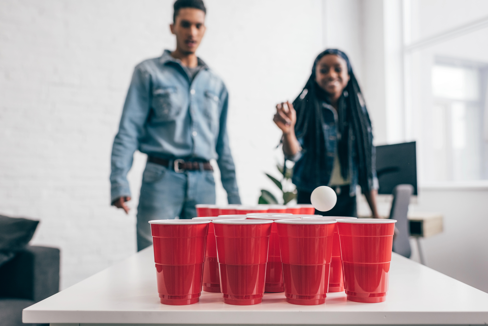 Two people play beer pong indoors. A ping pong ball is mid-air, aiming for a set of red cups on a white table. Both players are casually dressed and focused on the game. Bright natural light fills the room.