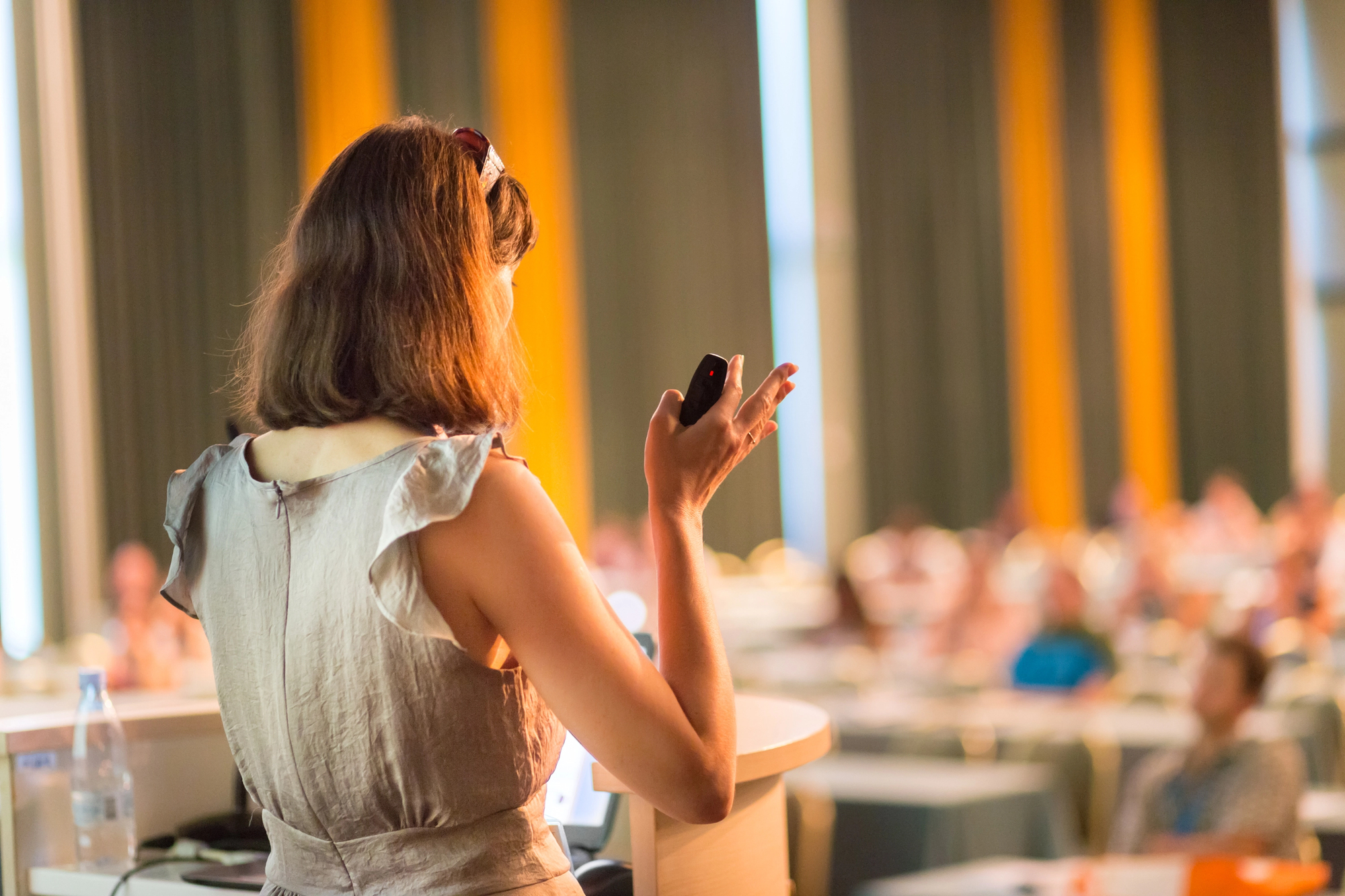 A woman in a beige dress stands at a podium with a microphone in hand, facing a blurred audience in a conference room. Vertical, yellow and gray curtains are visible in the background.