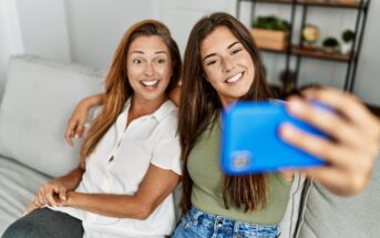 A woman and a young woman are sitting on a couch, smiling and taking a selfie with a blue smartphone. They appear relaxed and cheerful in a home setting with shelves in the background.