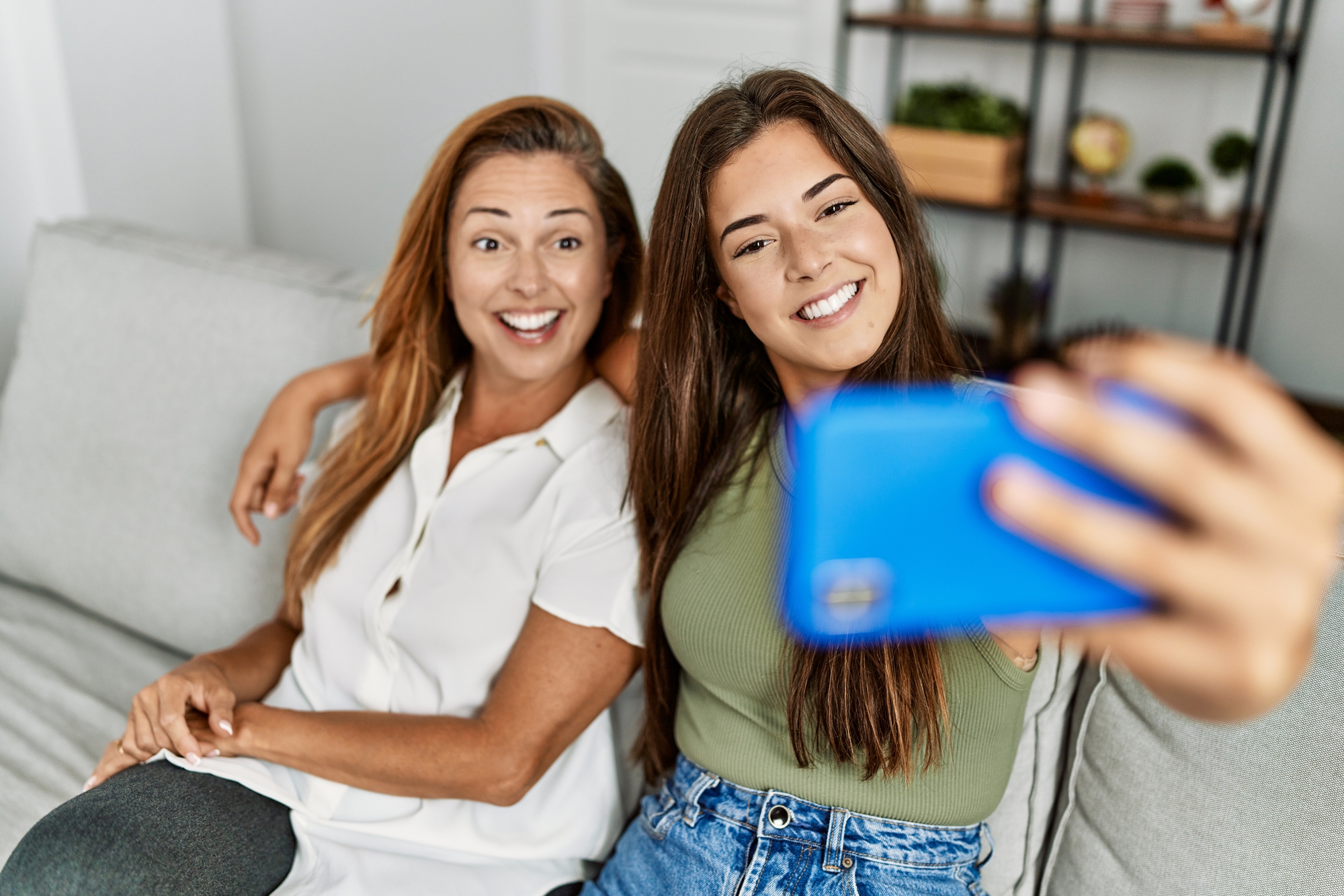 A woman and a young woman are sitting on a couch, smiling and taking a selfie with a blue smartphone. They appear relaxed and cheerful in a home setting with shelves in the background.