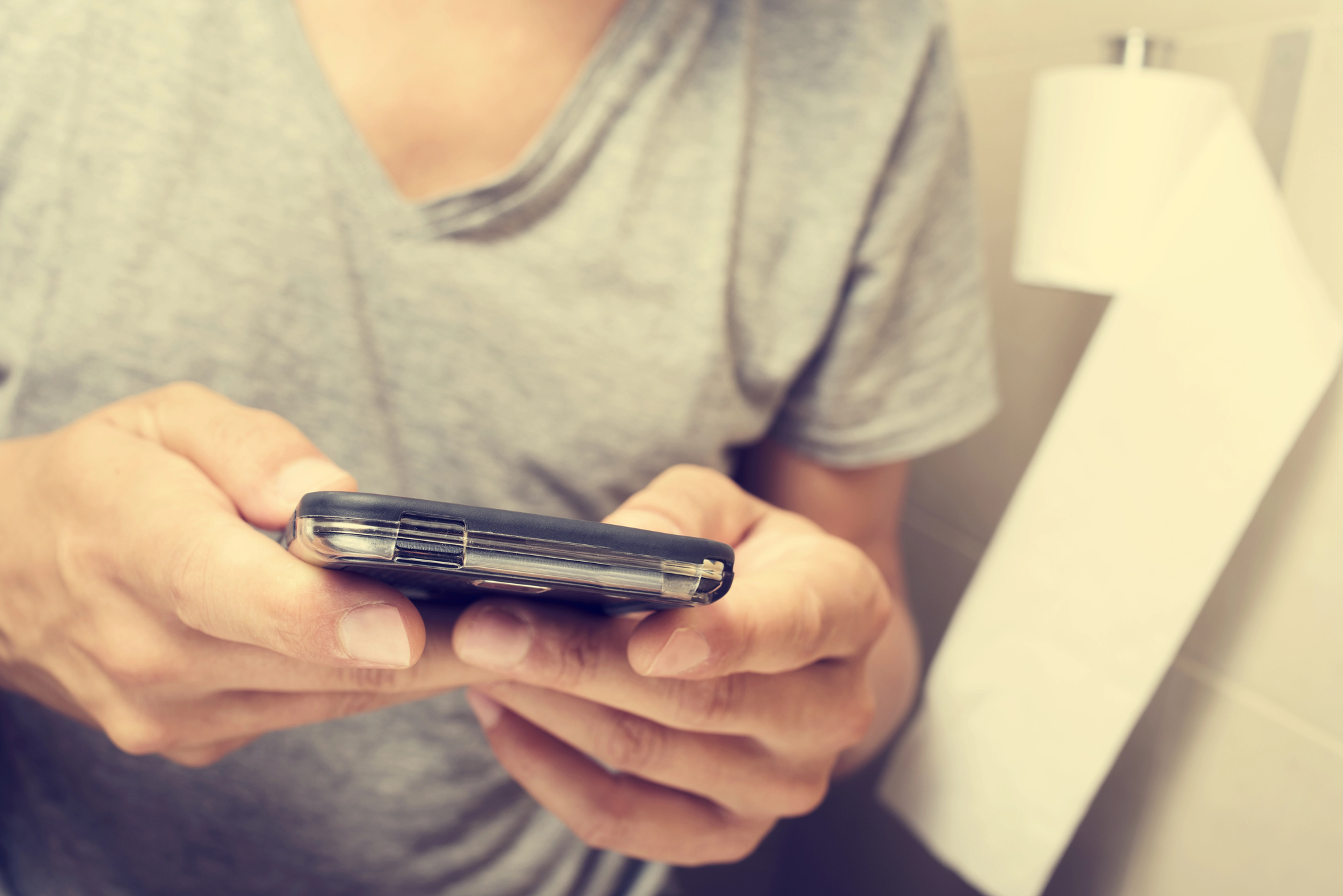 A person in a grey t-shirt is using a smartphone with both hands. In the background, there is a roll of toilet paper mounted on the wall. The setting appears to be a bathroom.