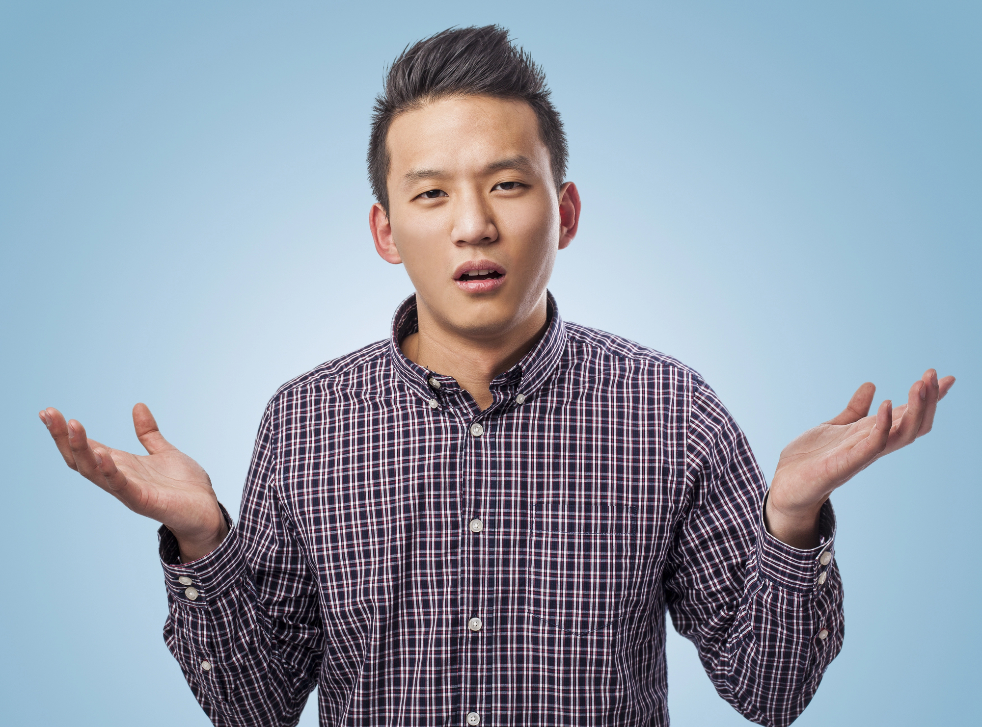 A person with short dark hair wearing a plaid shirt stands against a light blue background. They have a puzzled expression and their arms are raised with palms facing up, as if shrugging or questioning.