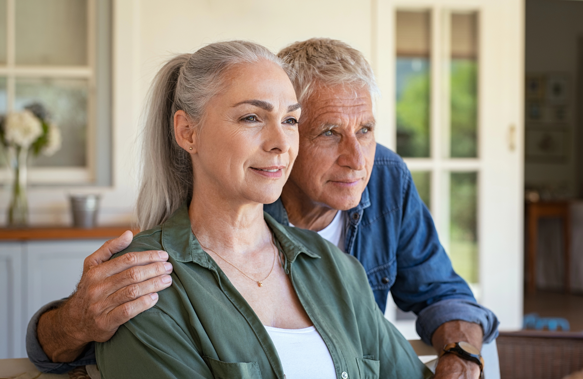 An older couple sits together; the woman with gray hair tied back and wearing a green blouse gazes ahead, while the man with short gray hair and a denim shirt looks in the same direction, resting his hand on her shoulder. They appear thoughtful and content.