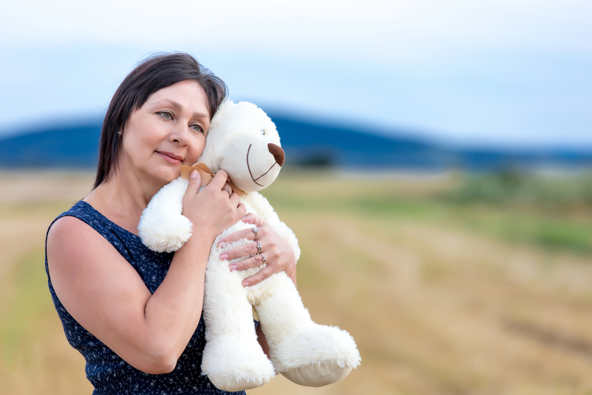 A woman with dark hair wearing a blue dress hugs a white teddy bear while standing on a rural path. The background features blurred fields and distant hills under a cloudy sky.