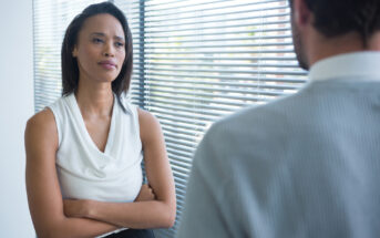 A woman with long, dark hair stands with her arms crossed, wearing a white sleeveless top, engaged in a serious conversation with a man in a light-colored shirt. They are in an office with blinds covering a large window.