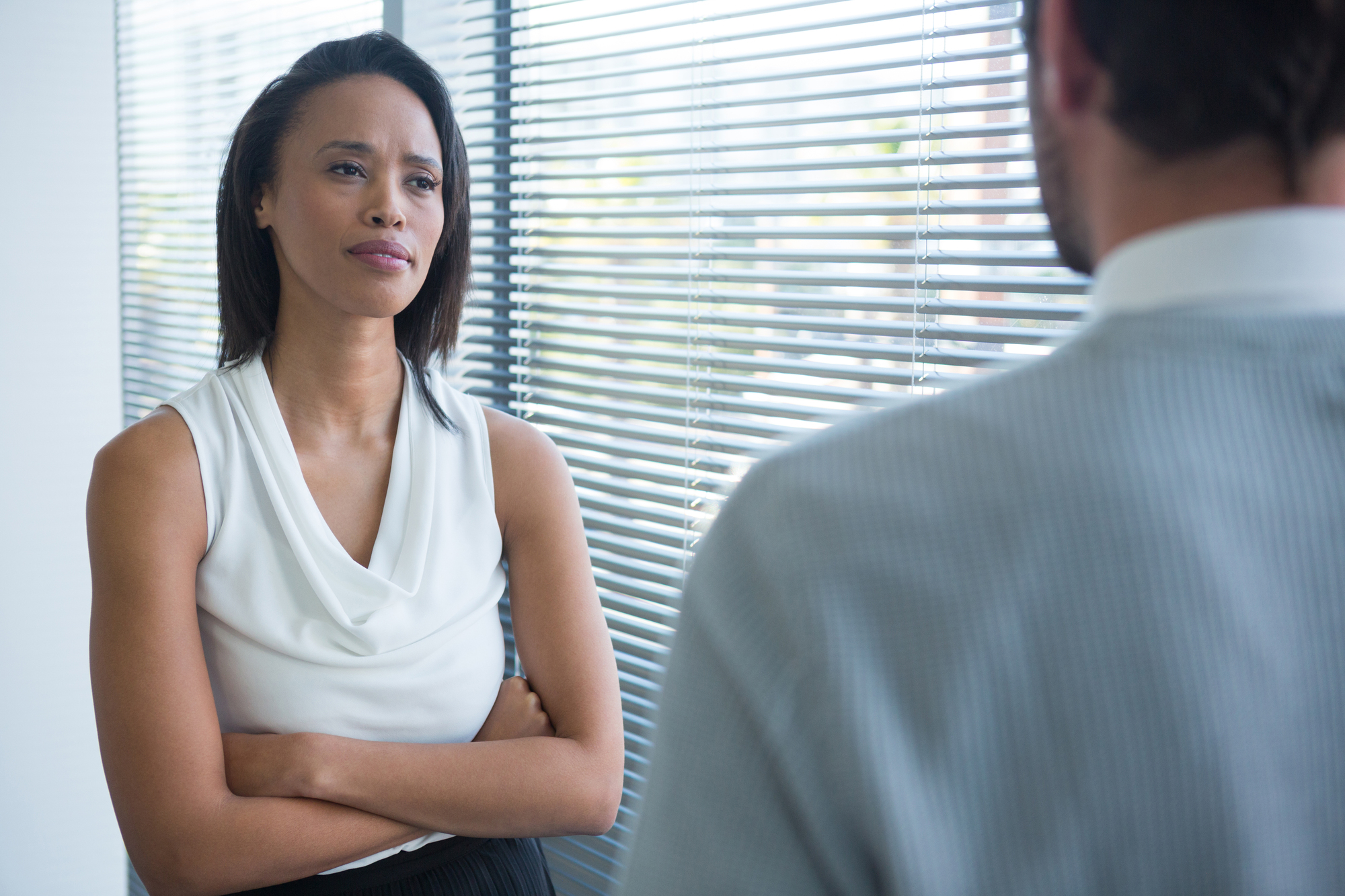 A woman with long, dark hair stands with her arms crossed, wearing a white sleeveless top, engaged in a serious conversation with a man in a light-colored shirt. They are in an office with blinds covering a large window.