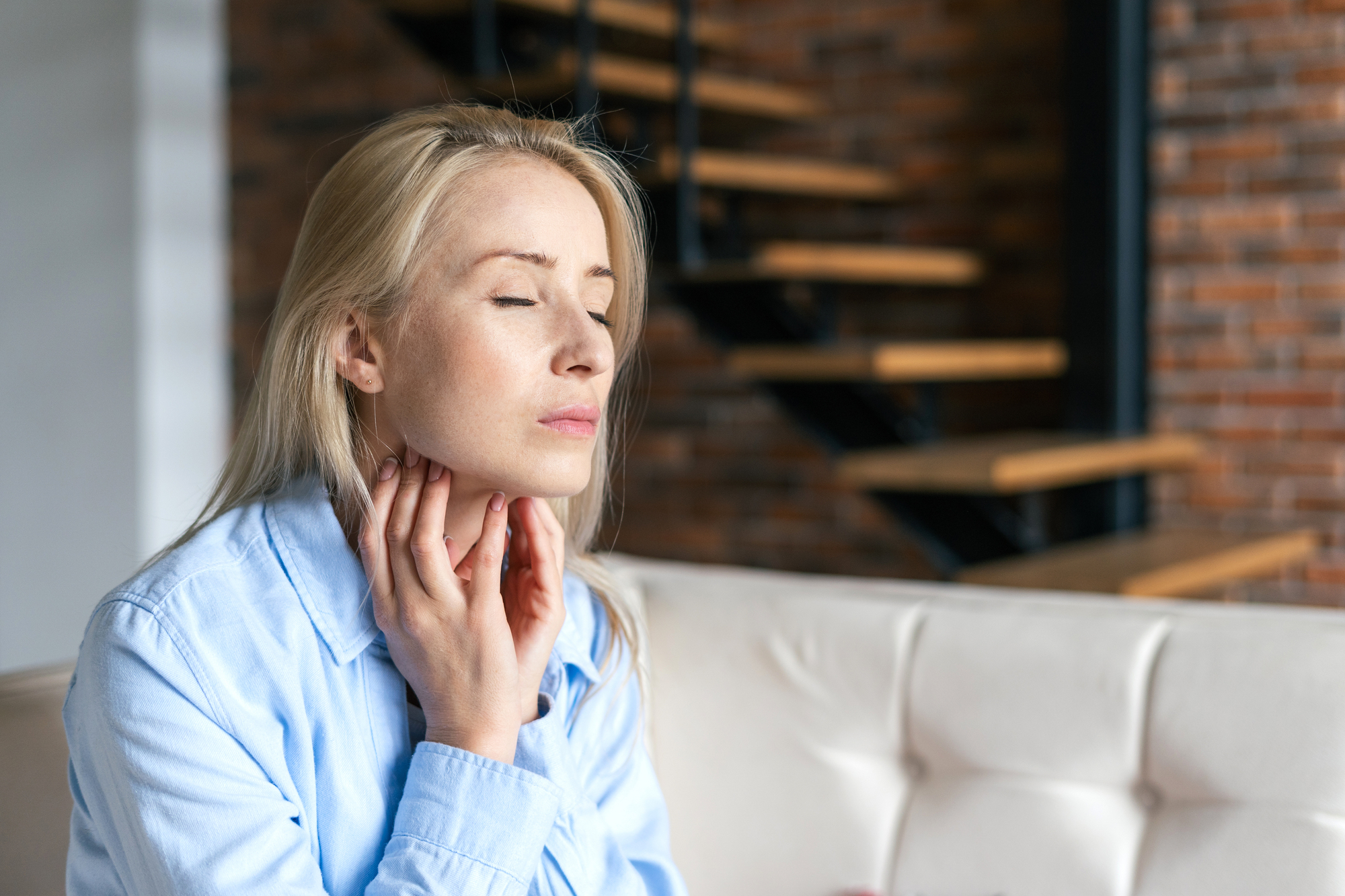 A woman with long blonde hair and a light blue shirt sits on a white couch with her eyes closed, touching her throat as if checking for a sore throat or discomfort. In the background, there are wooden stairs and a brick wall.