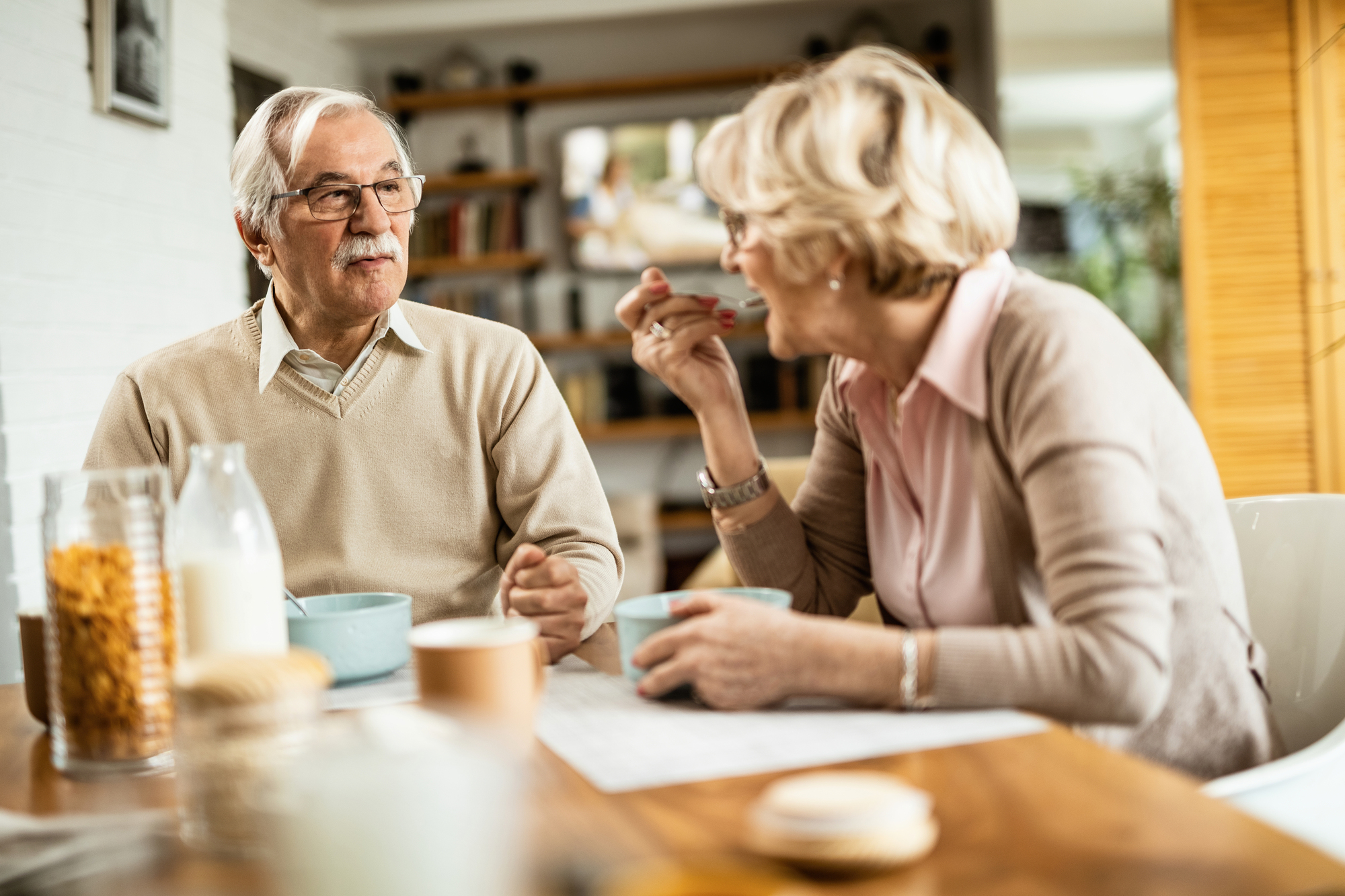 An elderly couple sits at a dining table enjoying breakfast. They are having a conversation, with bowls, a bottle of milk, and cereal on the table. Shelves with books and decor are visible in the background.