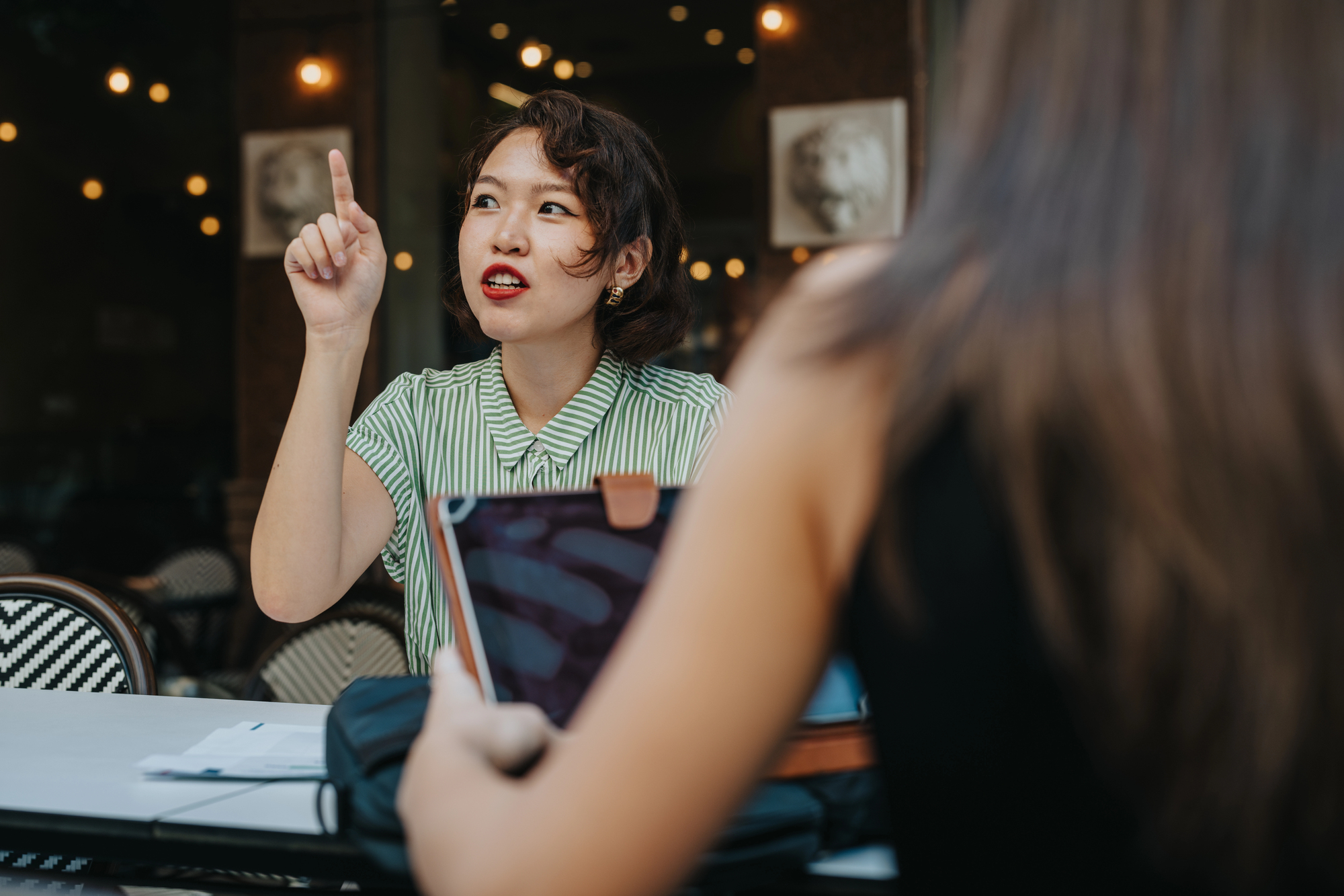 A woman in a green striped shirt gestures with her hand, sitting at an outdoor table. She is engaged in conversation with another person, whose back is visible. Warm string lights and blurred surroundings suggest a cafe setting.