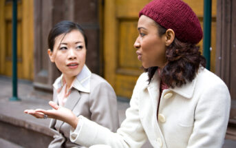 Two women in coats are sitting on steps in conversation. One has a red knit hat and gestures with her hand, while the other holds a coffee cup and listens attentively. Wooden doors are visible in the background.