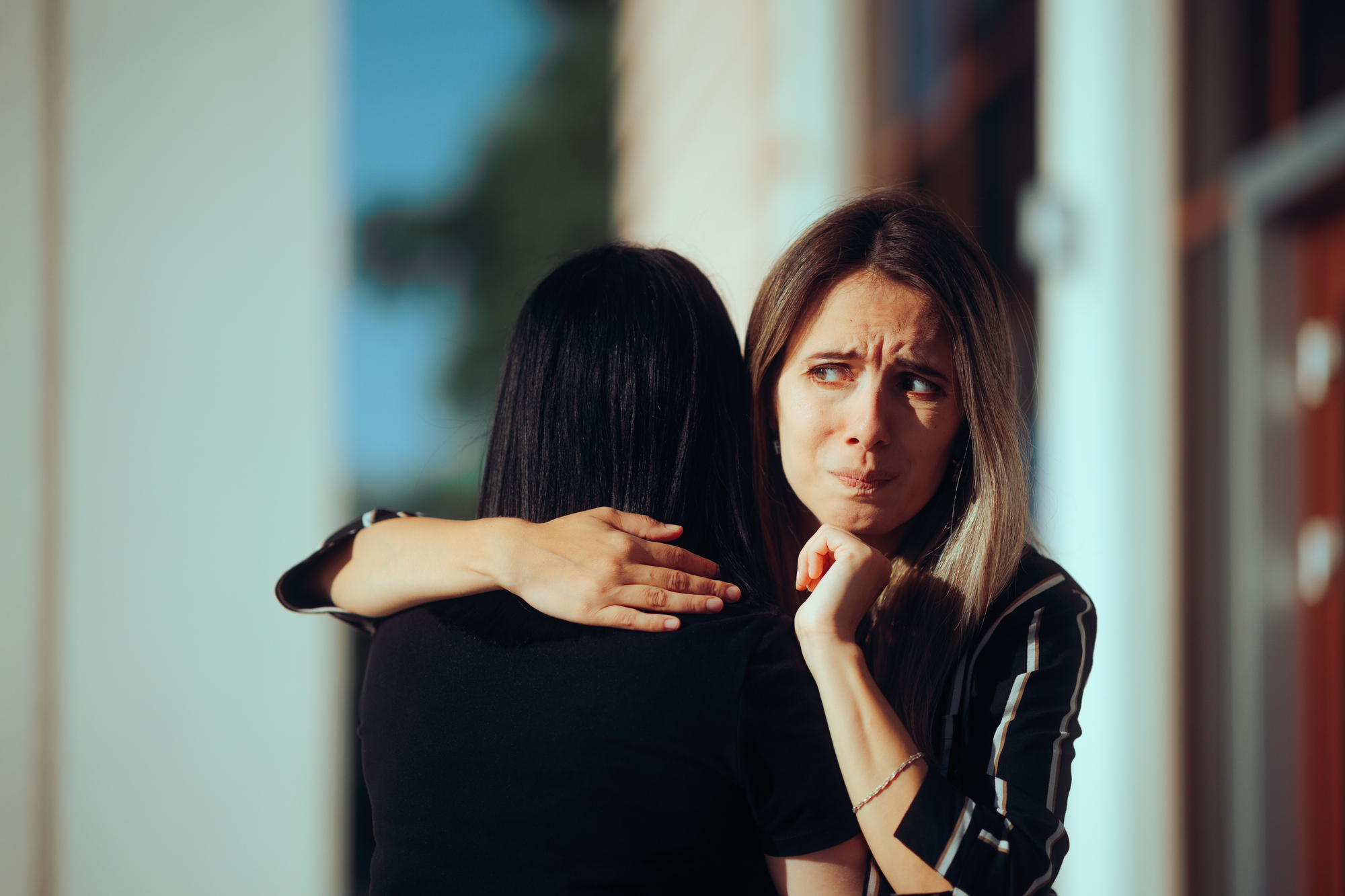 A woman with light-colored hair looks distressed while hugging another woman with dark hair. They stand outside near a building, and sunlight casts shadows around them.