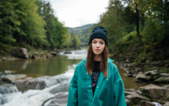 A young woman stands by a river surrounded by greenery. She is wearing a black beanie, black shirt, and a teal jacket. The river flows over rocks, and trees line the banks, with hills in the distance. The scene is tranquil and natural.