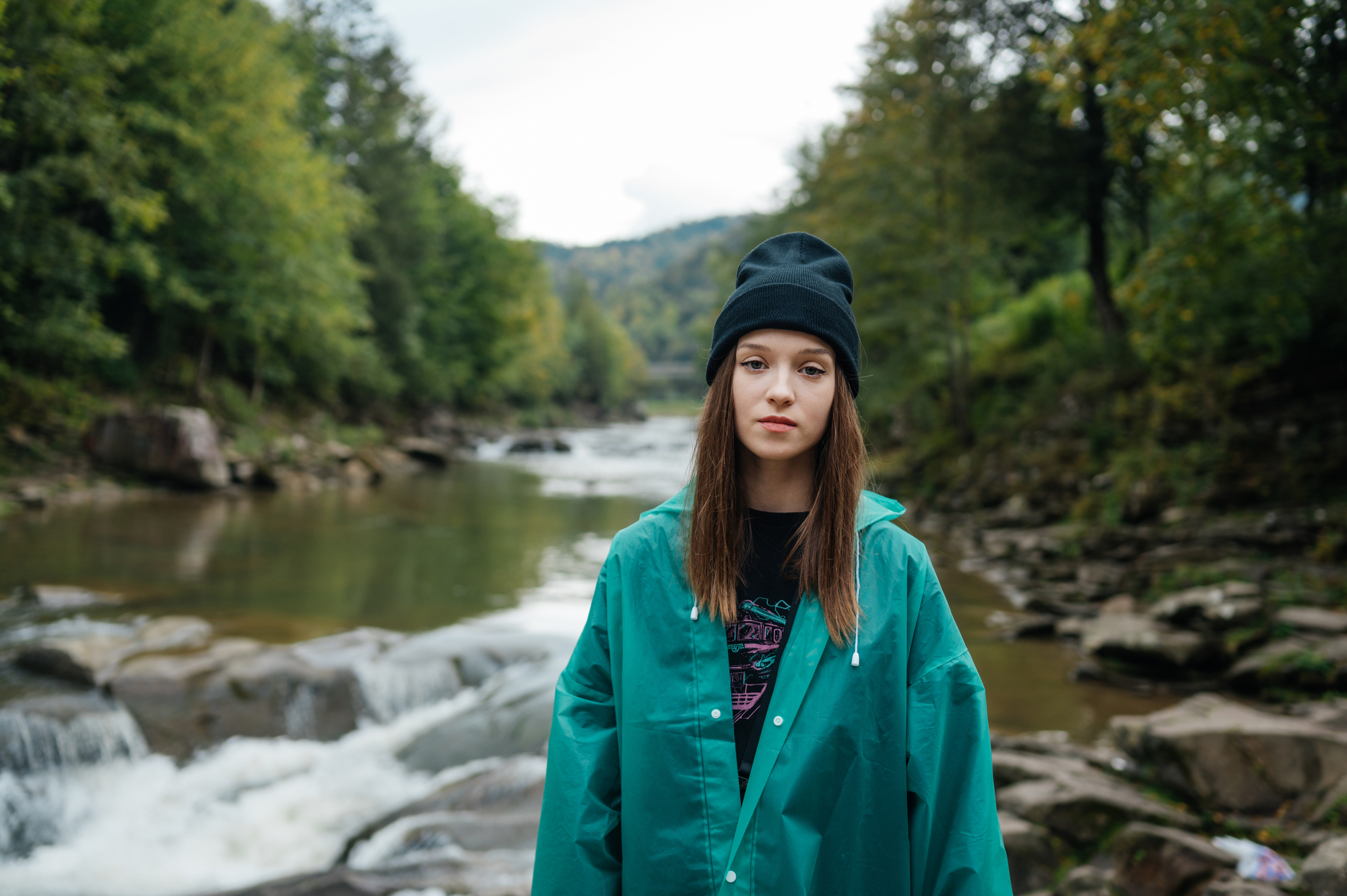 A young woman stands by a river surrounded by greenery. She is wearing a black beanie, black shirt, and a teal jacket. The river flows over rocks, and trees line the banks, with hills in the distance. The scene is tranquil and natural.