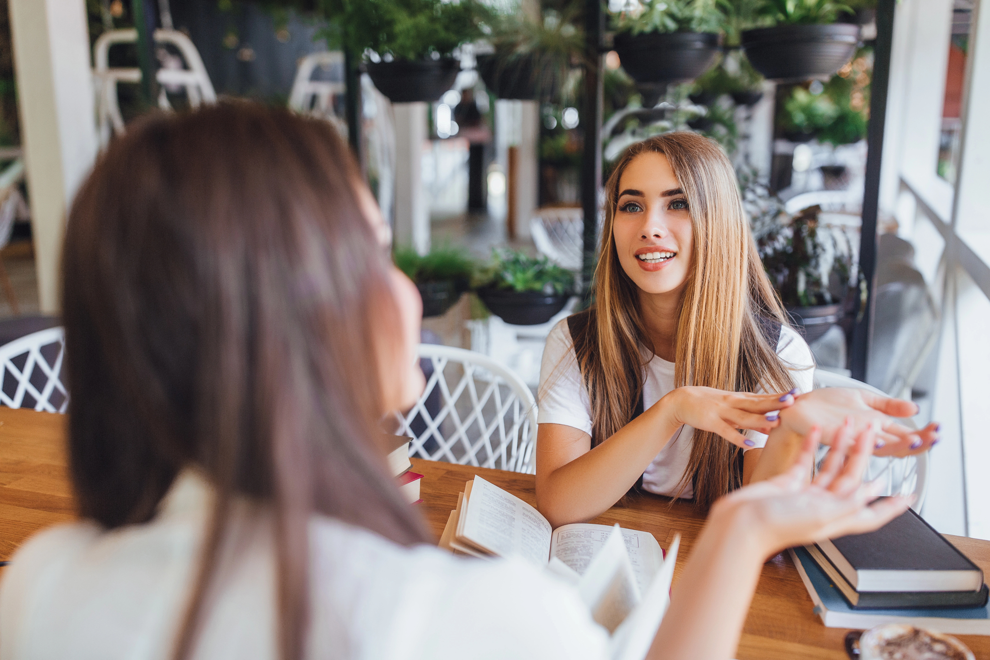 Two women are sitting at a wooden table in a cozy indoor setting with lots of plants. They are engaged in conversation, with an open book on the table. The woman facing the camera has long hair and is smiling while gesturing with her hands.