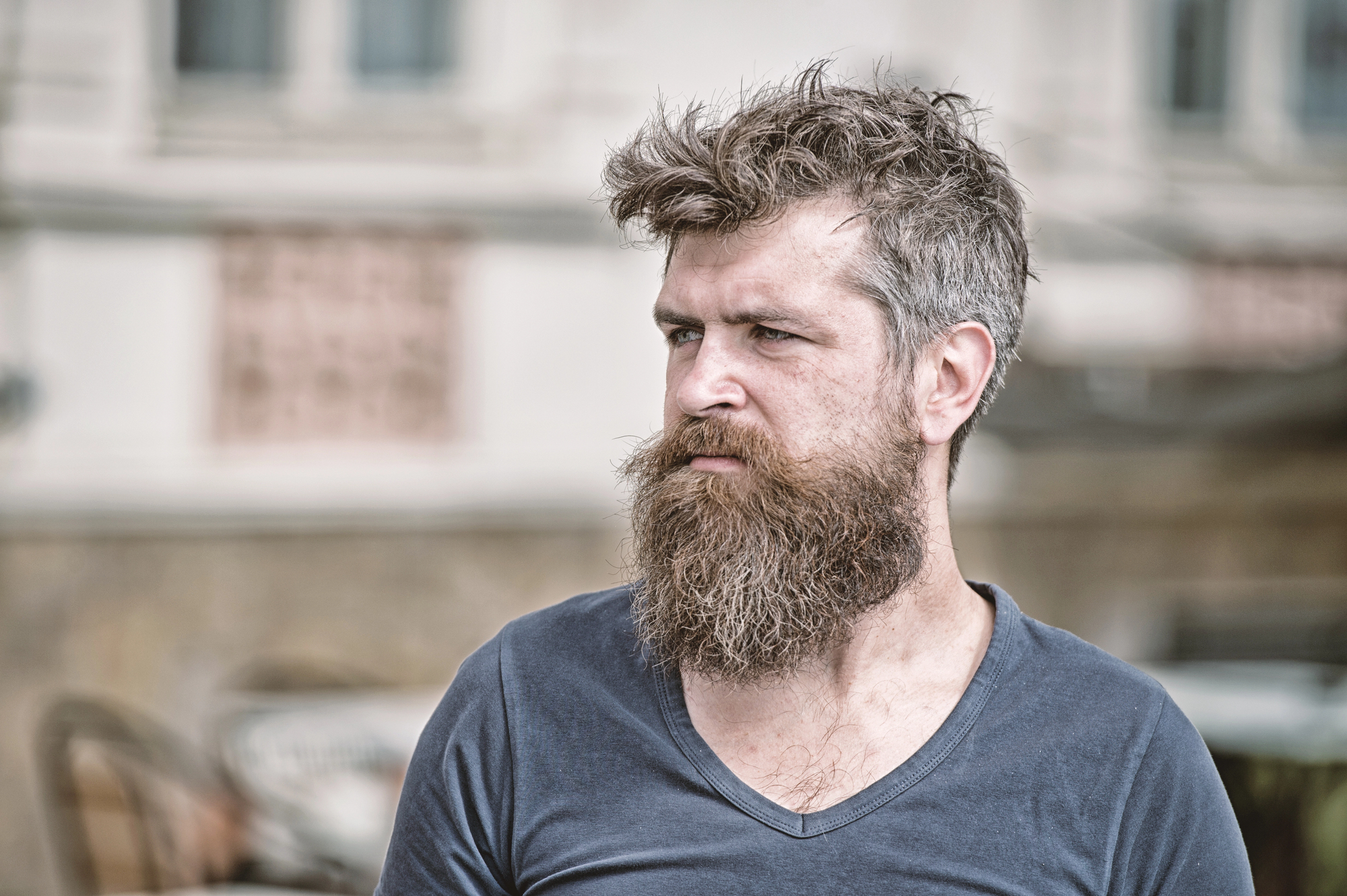 A man with a thick beard and tousled hair looks to the side. He is wearing a V-neck shirt and is outdoors, with blurred buildings and chairs in the background.