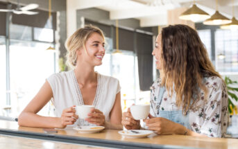 Two women sitting at a cafe table, smiling and holding white mugs. They appear engaged in conversation, with a bright, modern interior featuring large windows and hanging lights in the background.