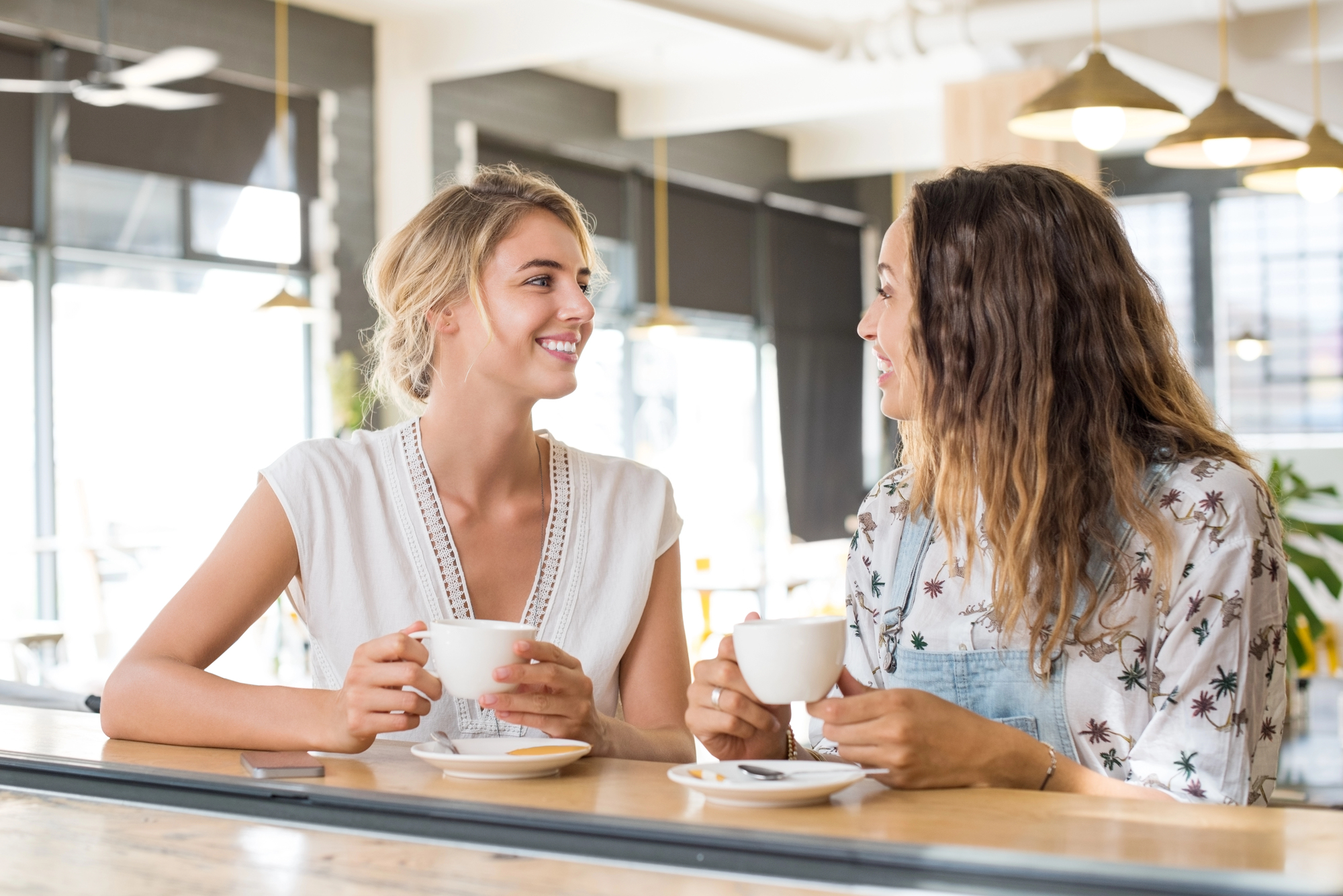 Two women sitting at a cafe table, smiling and holding white mugs. They appear engaged in conversation, with a bright, modern interior featuring large windows and hanging lights in the background.