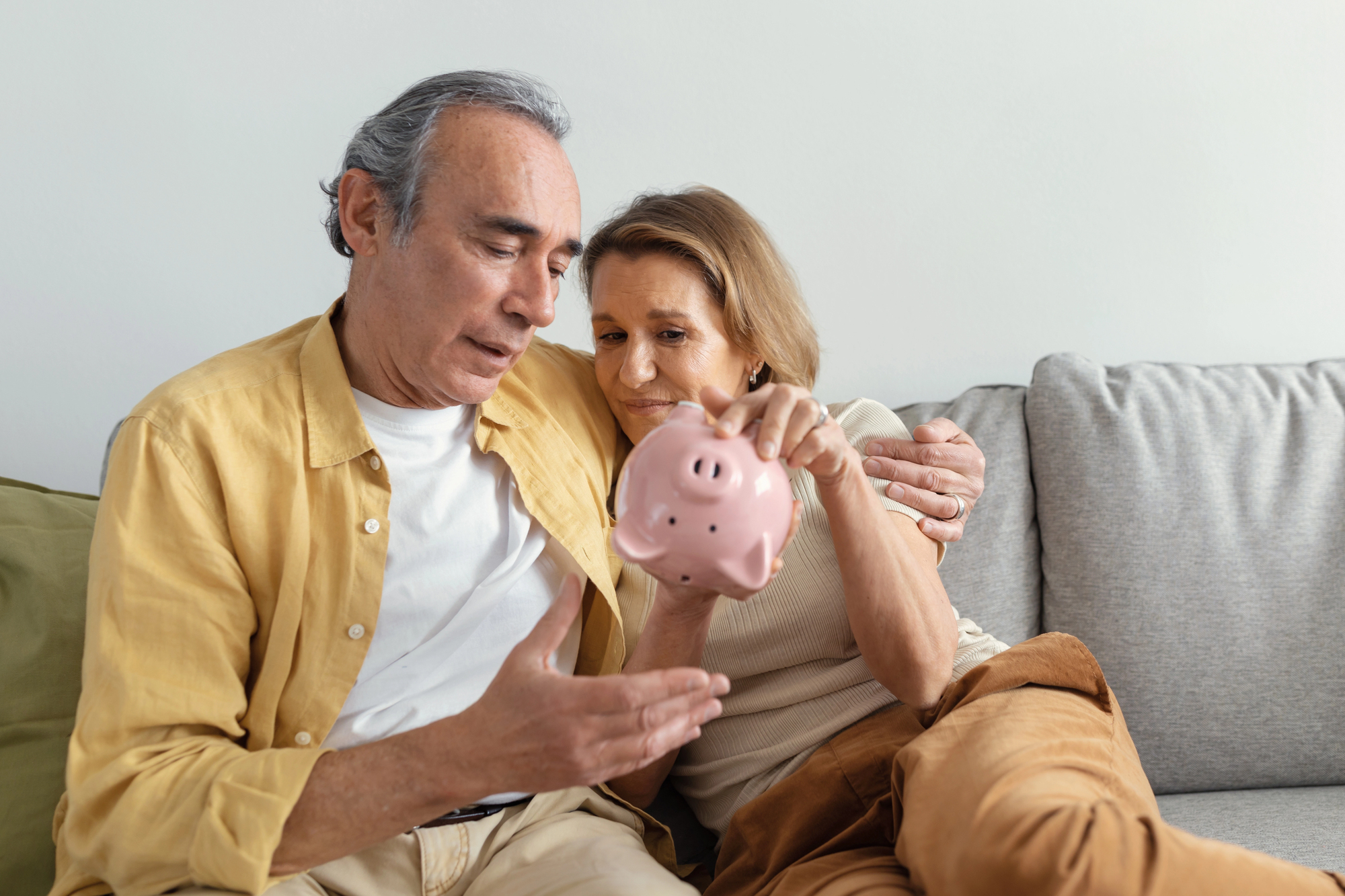 An older couple sits on a sofa, closely examining a pink piggy bank. The man, wearing a yellow shirt, gestures with one hand, while the woman, in a beige sweater, looks attentively at the bank. Both appear thoughtful and engaged.