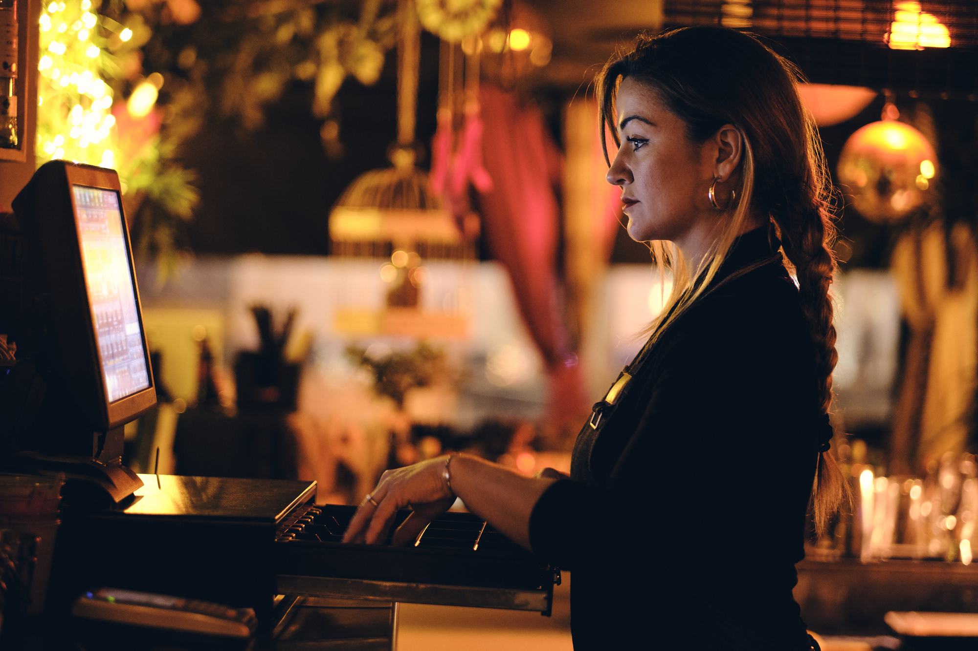 A woman with a braid works at a computer in a warmly lit, cozy environment. She is focused on the screen, with various decorative elements like plants and lights visible around her.