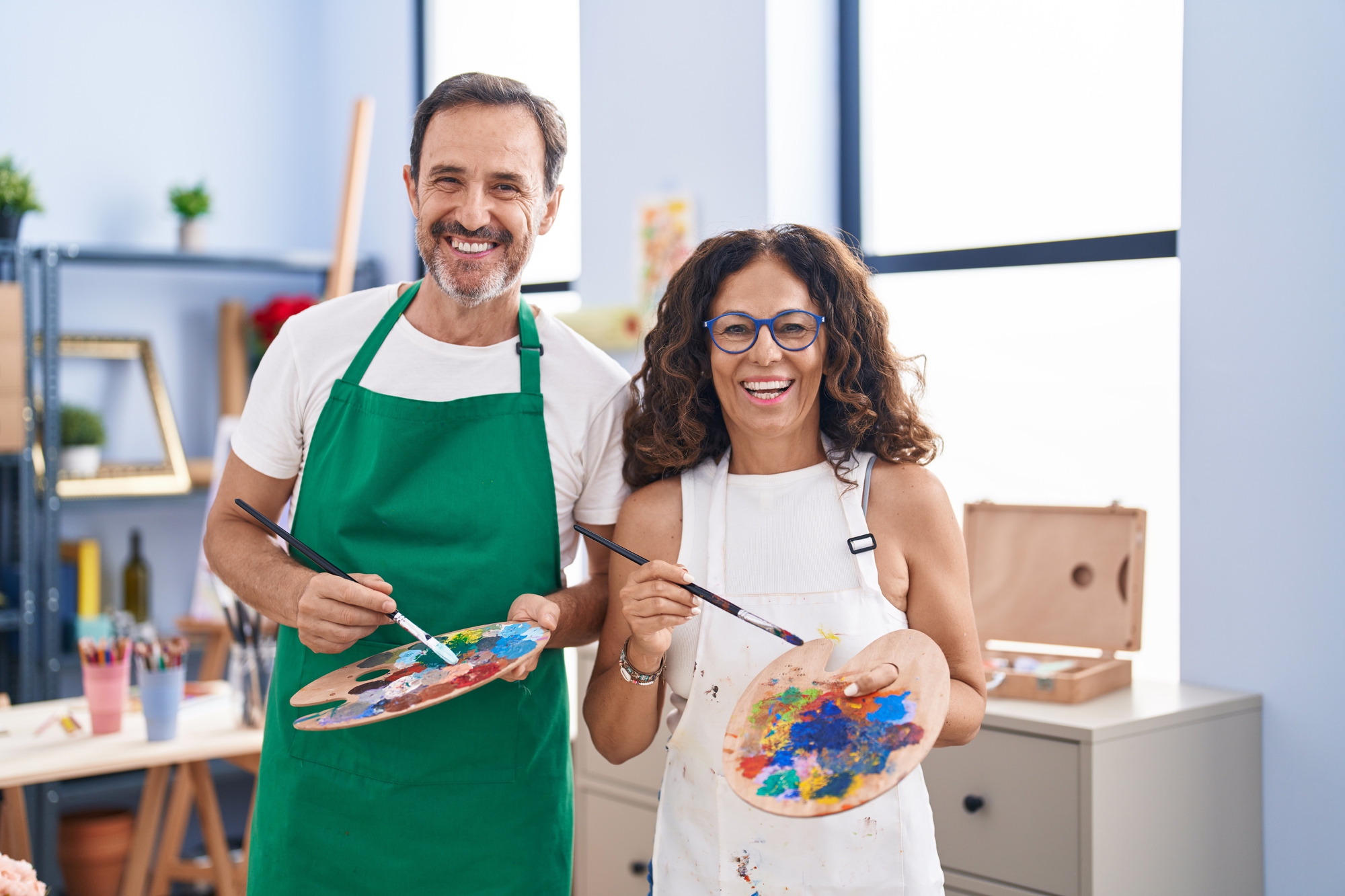 A man and woman are smiling while holding paint palettes and brushes. They are wearing aprons, standing in a brightly lit art studio with shelves and art supplies in the background. The atmosphere is cheerful and creative.