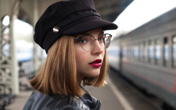 A person with shoulder-length hair, wearing glasses, a black hat, and a leather jacket stands on a train platform with a blurred train in the background. They look confidently towards the camera.