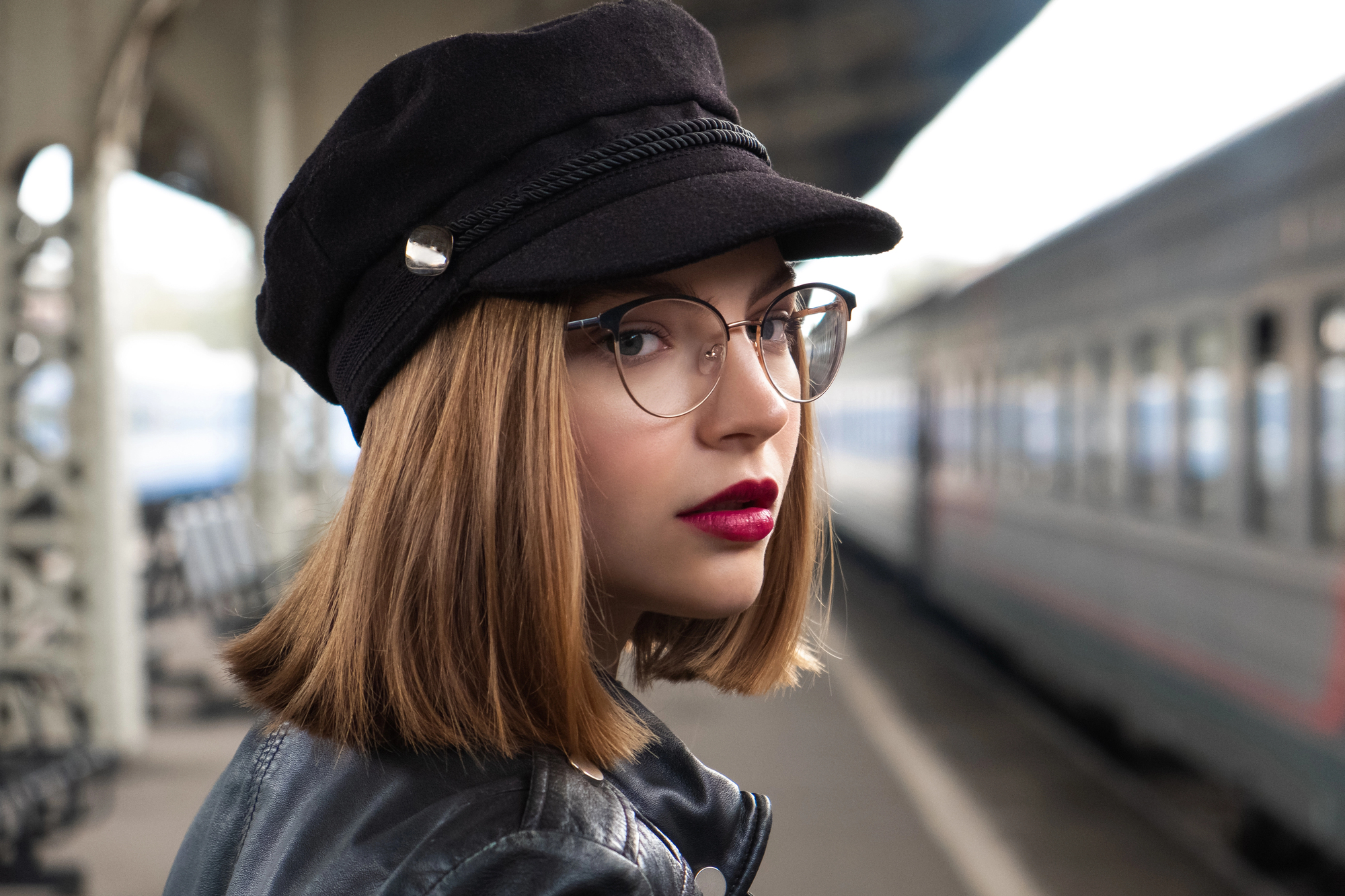 A person with shoulder-length hair, wearing glasses, a black hat, and a leather jacket stands on a train platform with a blurred train in the background. They look confidently towards the camera.