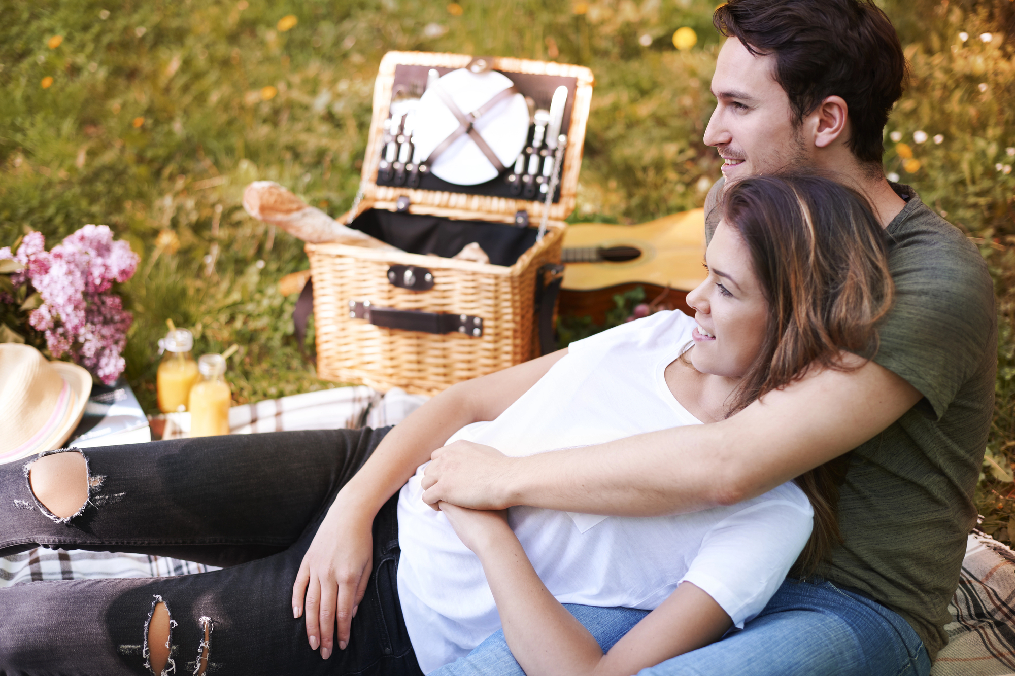 A couple relaxes on a picnic blanket in a grassy field, cuddling and smiling. Behind them is a wicker picnic basket with utensils exposed. Two glasses of juice and a bouquet of flowers are in view. The atmosphere is peaceful and sunny.