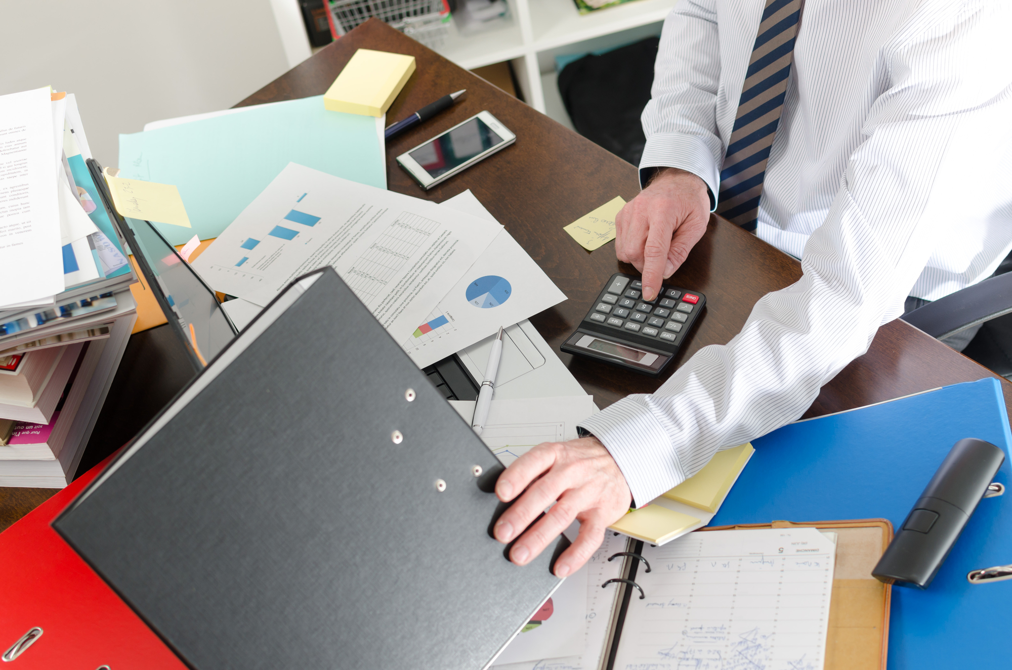 A person wearing a striped tie uses a calculator at a cluttered desk. The desk has documents with charts, a smartphone, colorful binders, and sticky notes. The person appears to be engaged in office work or financial calculations.