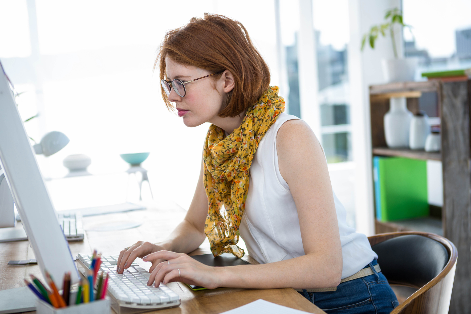 A woman with short red hair and glasses, wearing a white sleeveless top and a yellow scarf, is sitting at a desk, typing on a keyboard in a bright, modern office space. Various office supplies are on the desk.